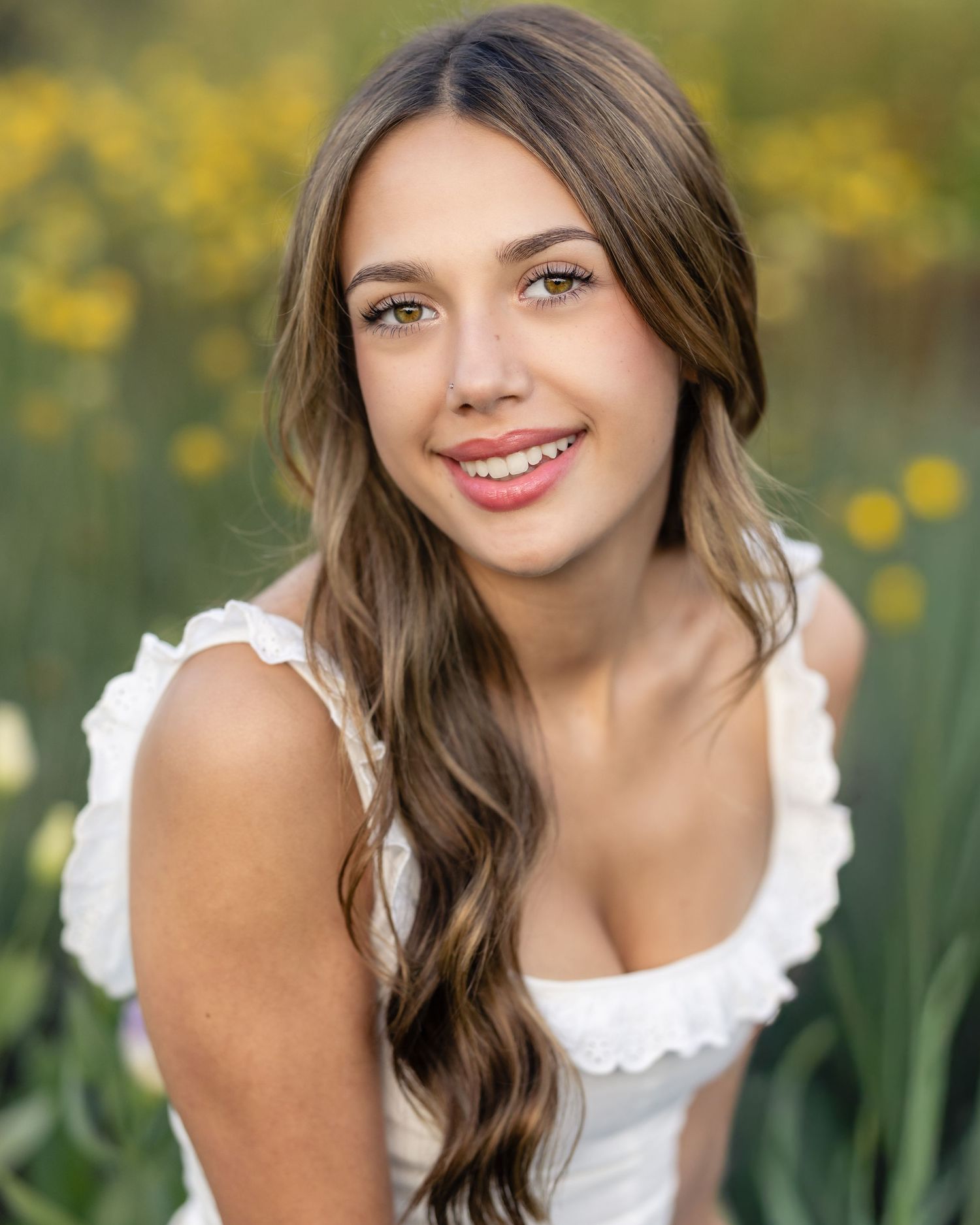 A young woman with long brown hair and a white dress smiles while surrounded by yellow flowers.