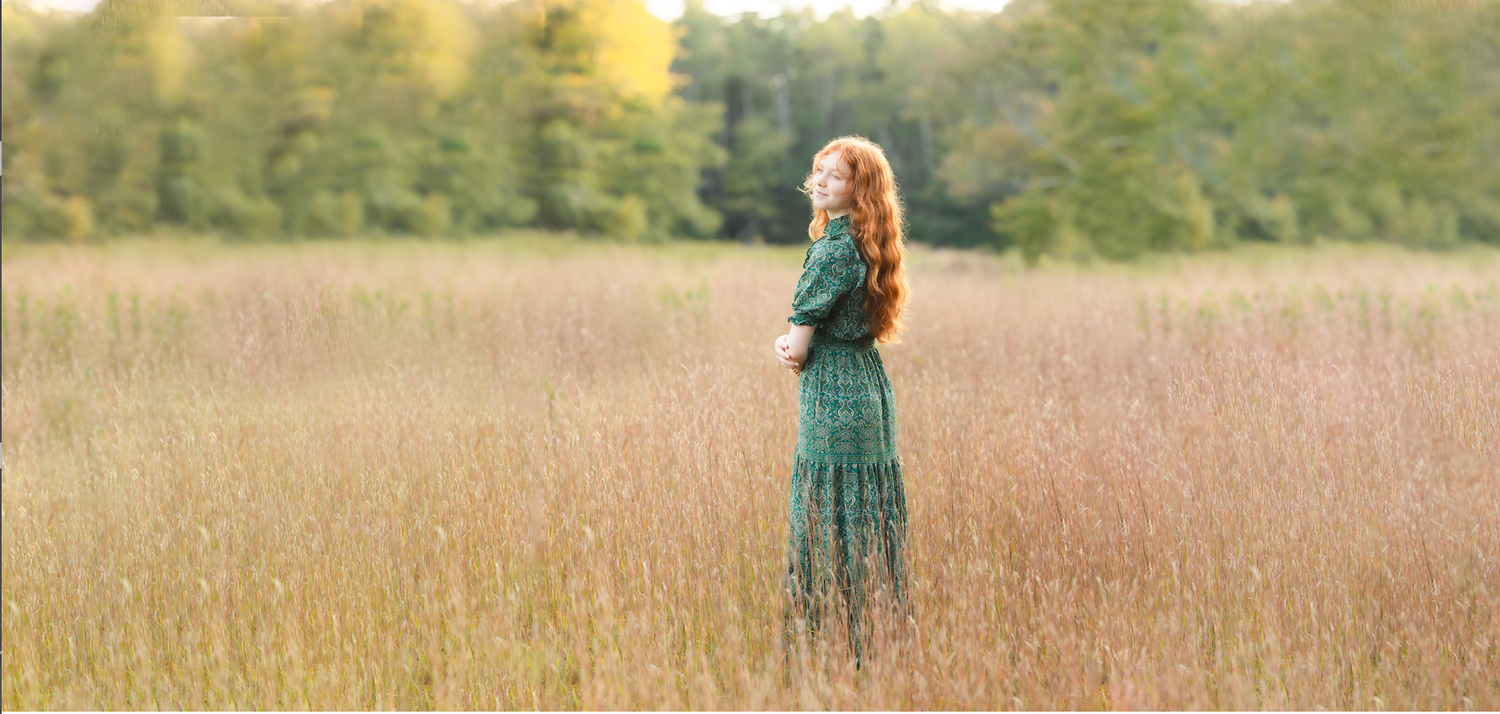 A woman in a green floral dress stands in a golden field, surrounded by lush trees in the background.
