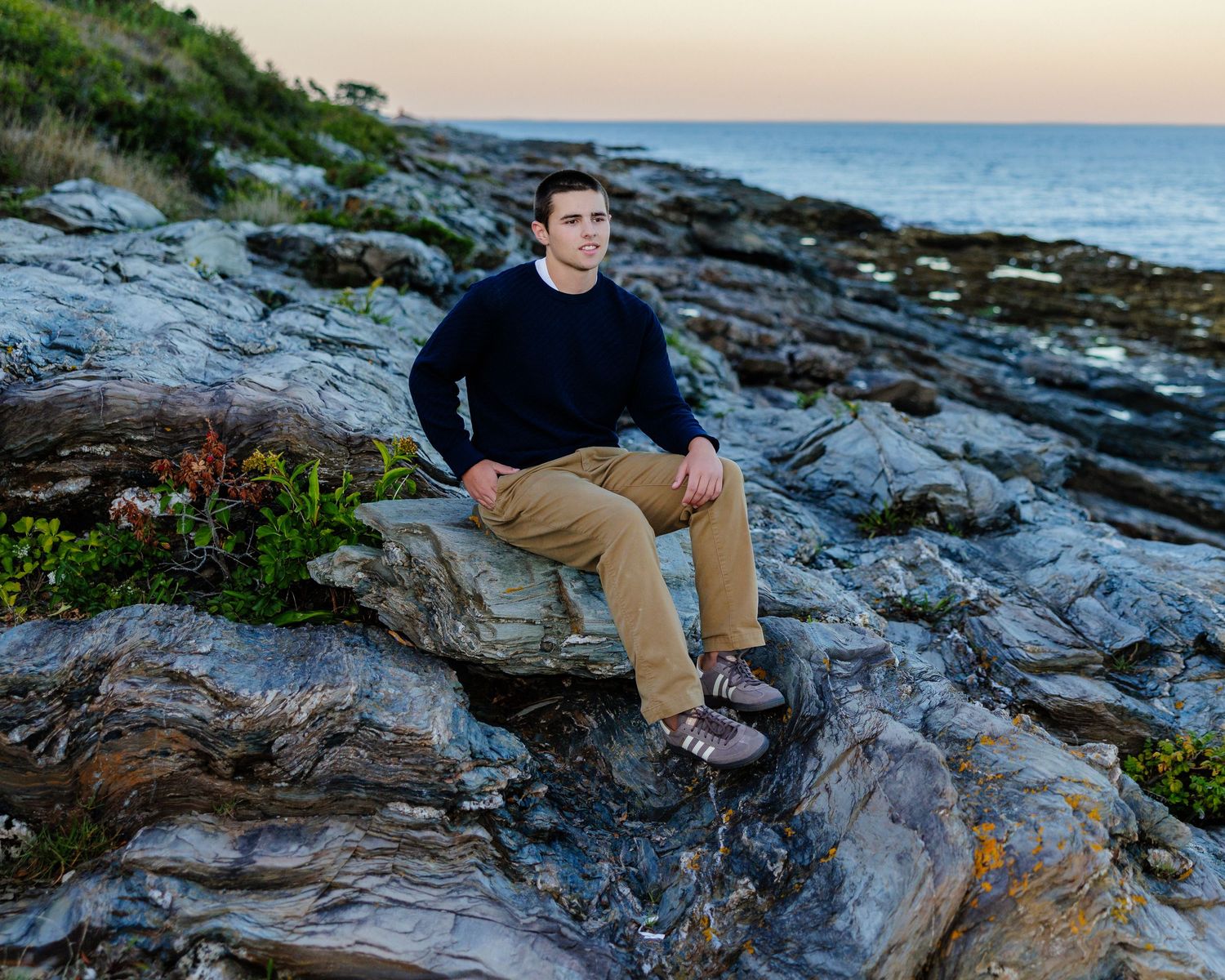 Young man in navy sweater and khaki pants sitting on rocky coastal shoreline at sunset.