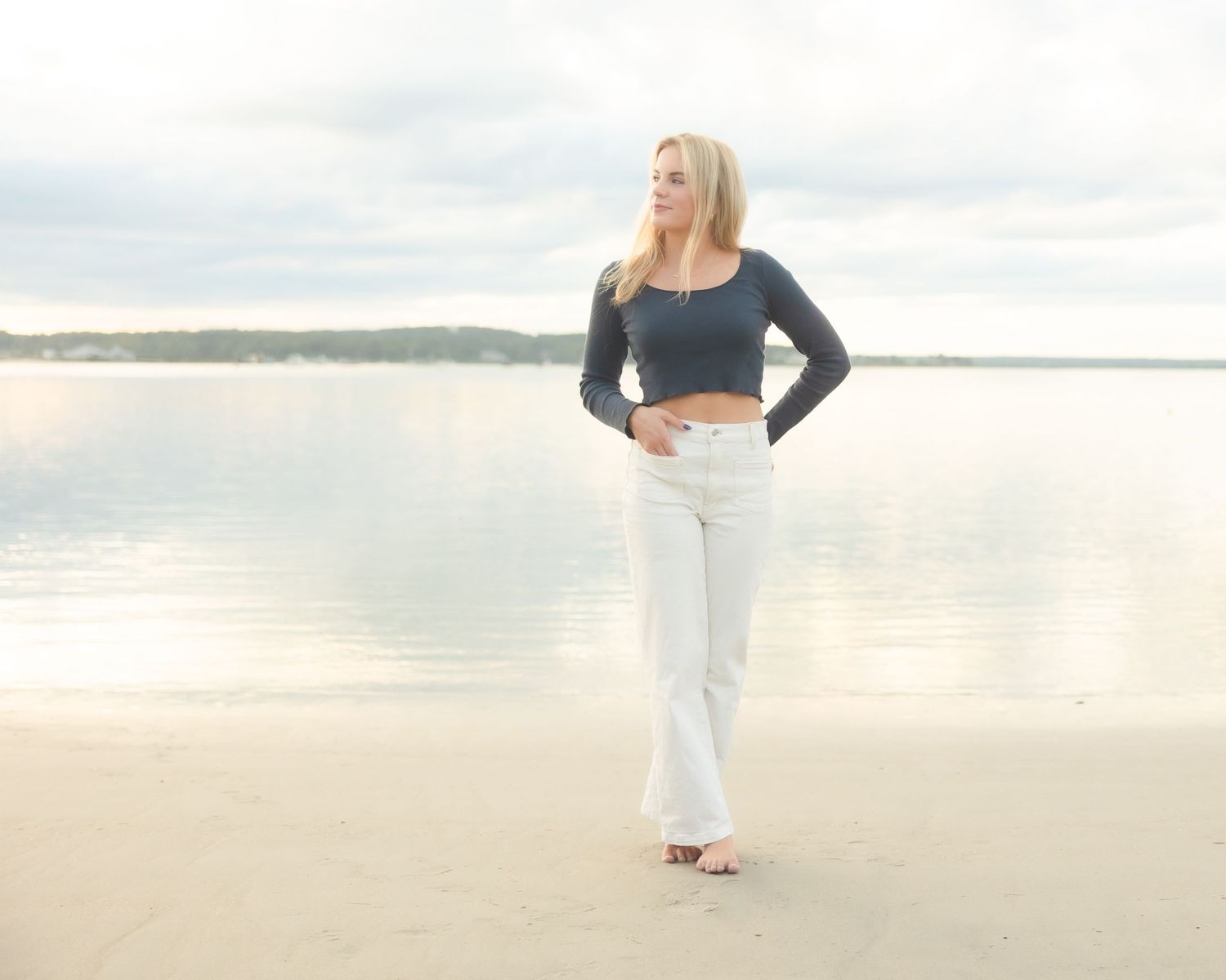 Blonde woman in crop top and white flare jeans standing barefoot on sandy beach with calm water backdrop.