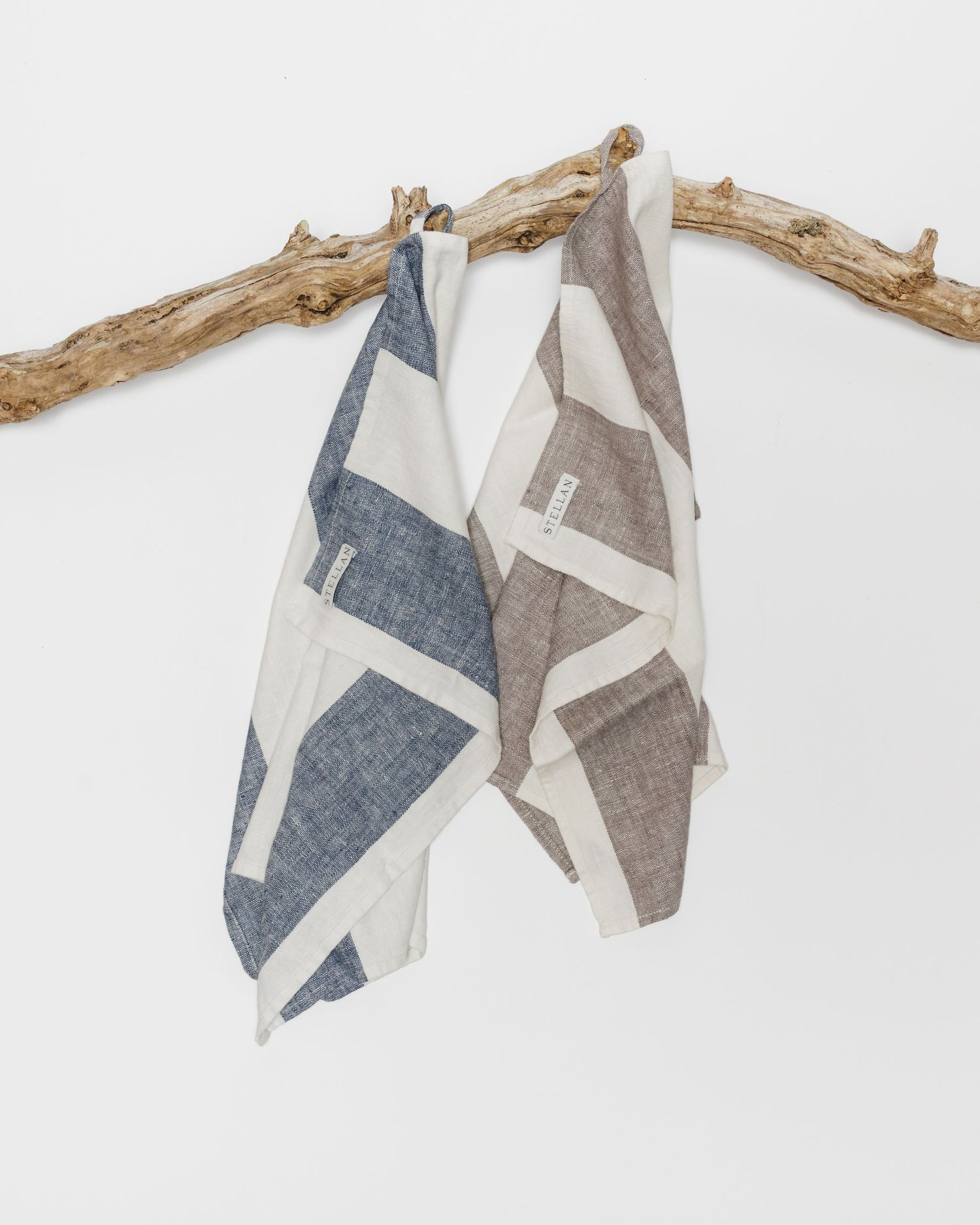Two geometric patterned linen tea towels in blue and brown hanging from a rustic driftwood branch.
