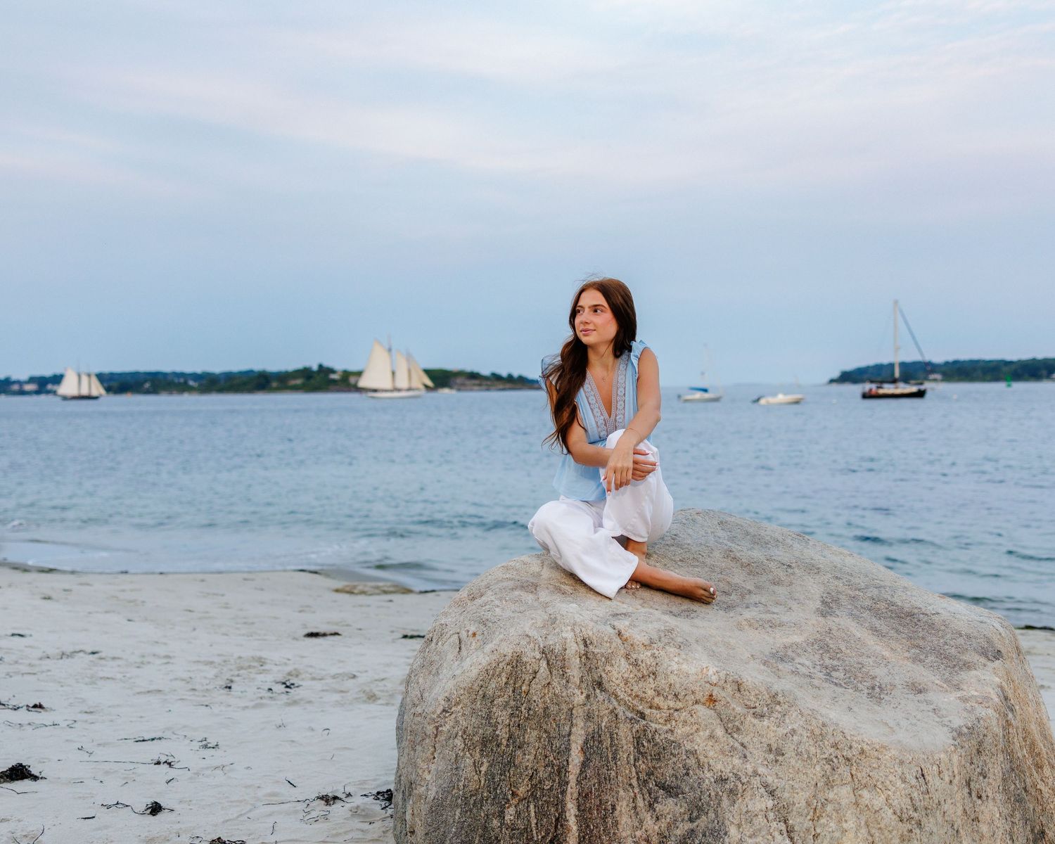 A woman in white outfit sits on a rock by a calm seaside beach with sailboats in the background.