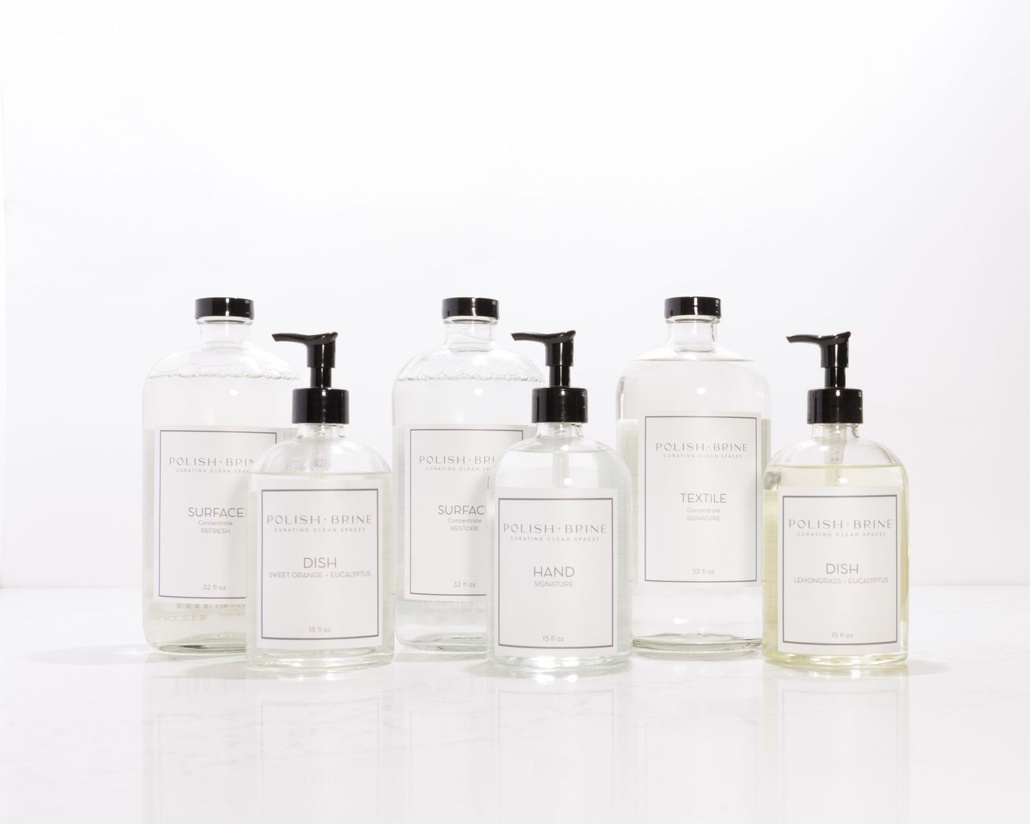 Collection of clear glass pump bottles with white labels arranged in varying sizes on a white surface.