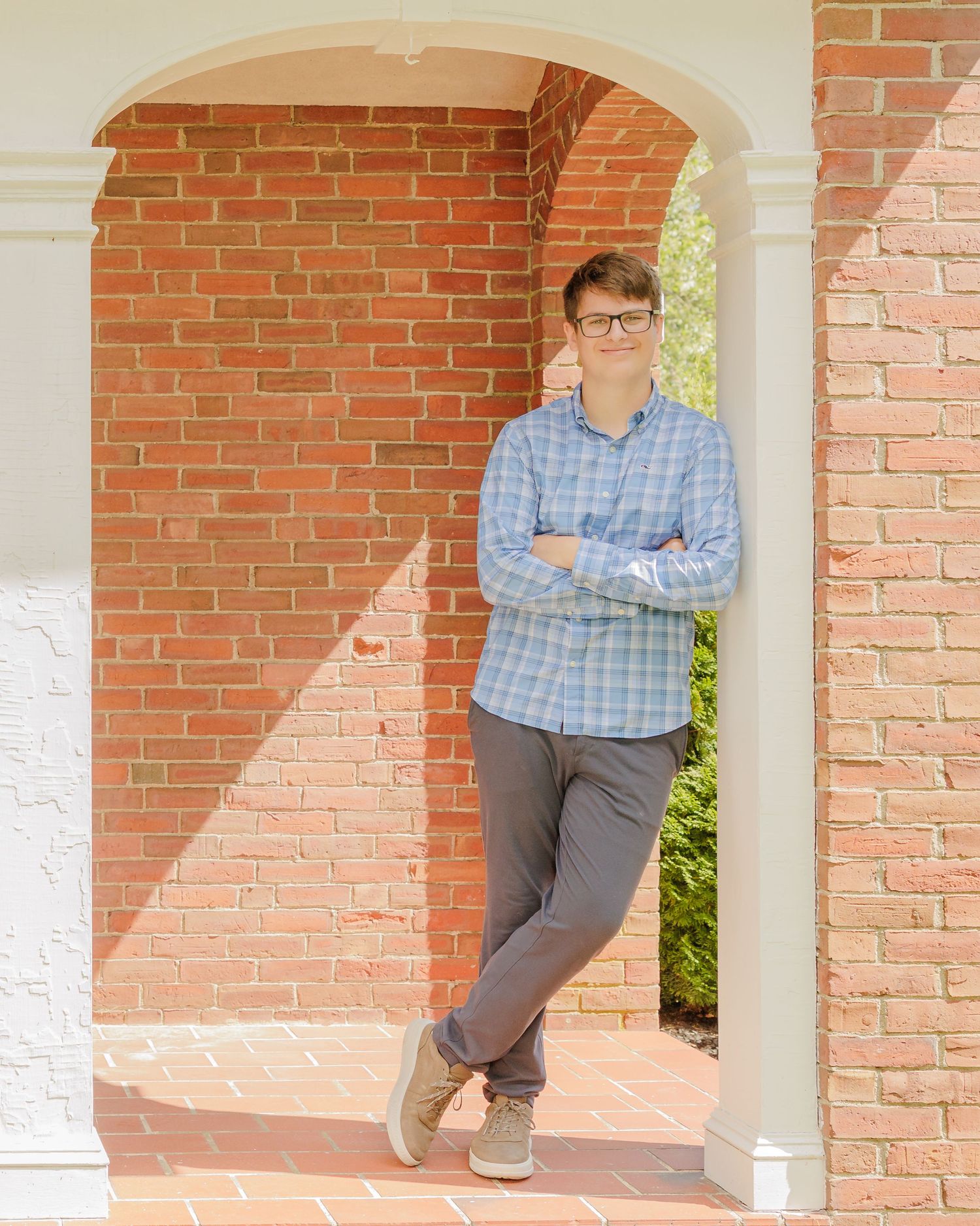 Young man in plaid shirt and glasses leaning against brick archway column with arms crossed.