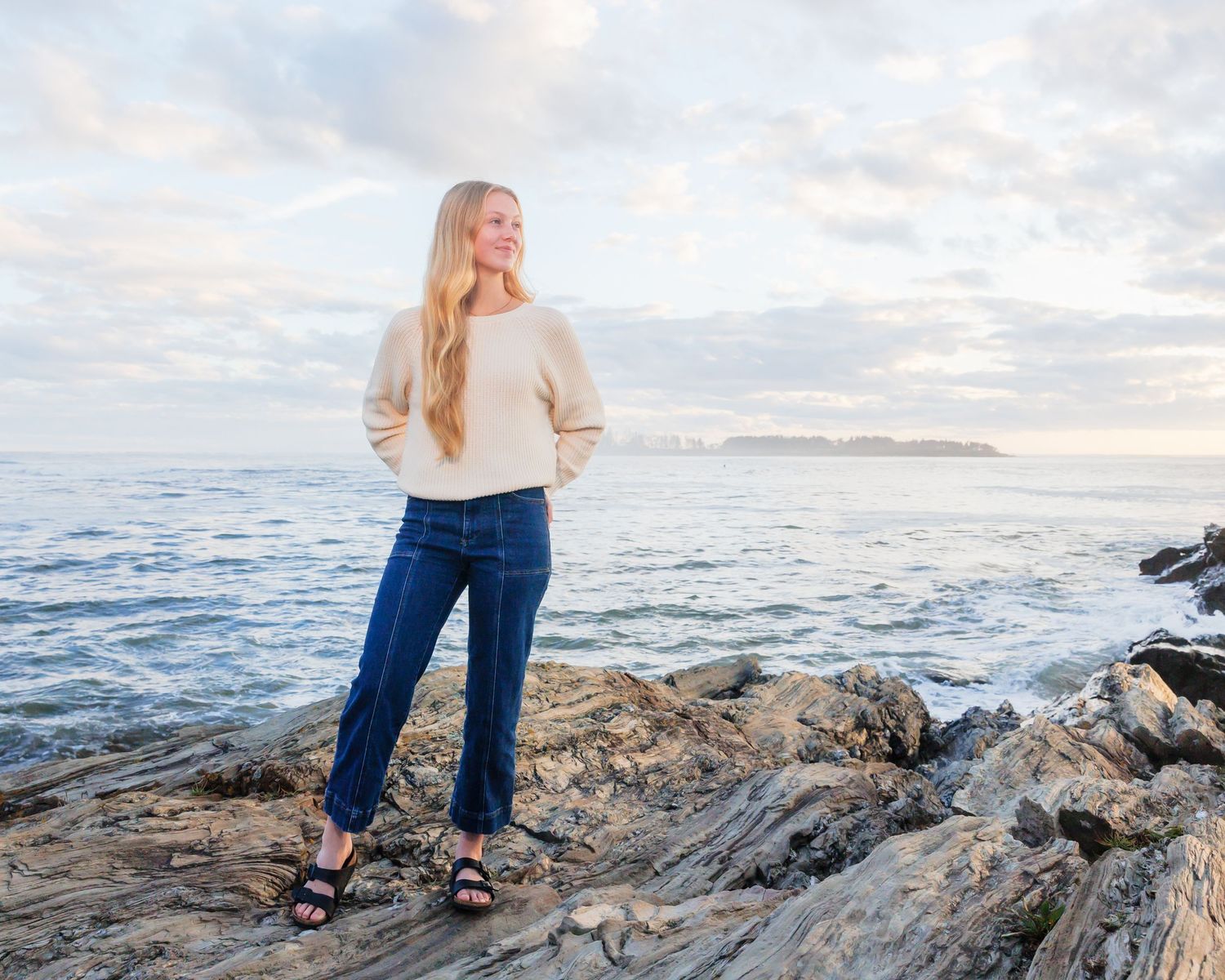Southern Maine Senior Portrait Photographer - Person in beige sweater stands on coastal rocks overlooking ocean at sunset.