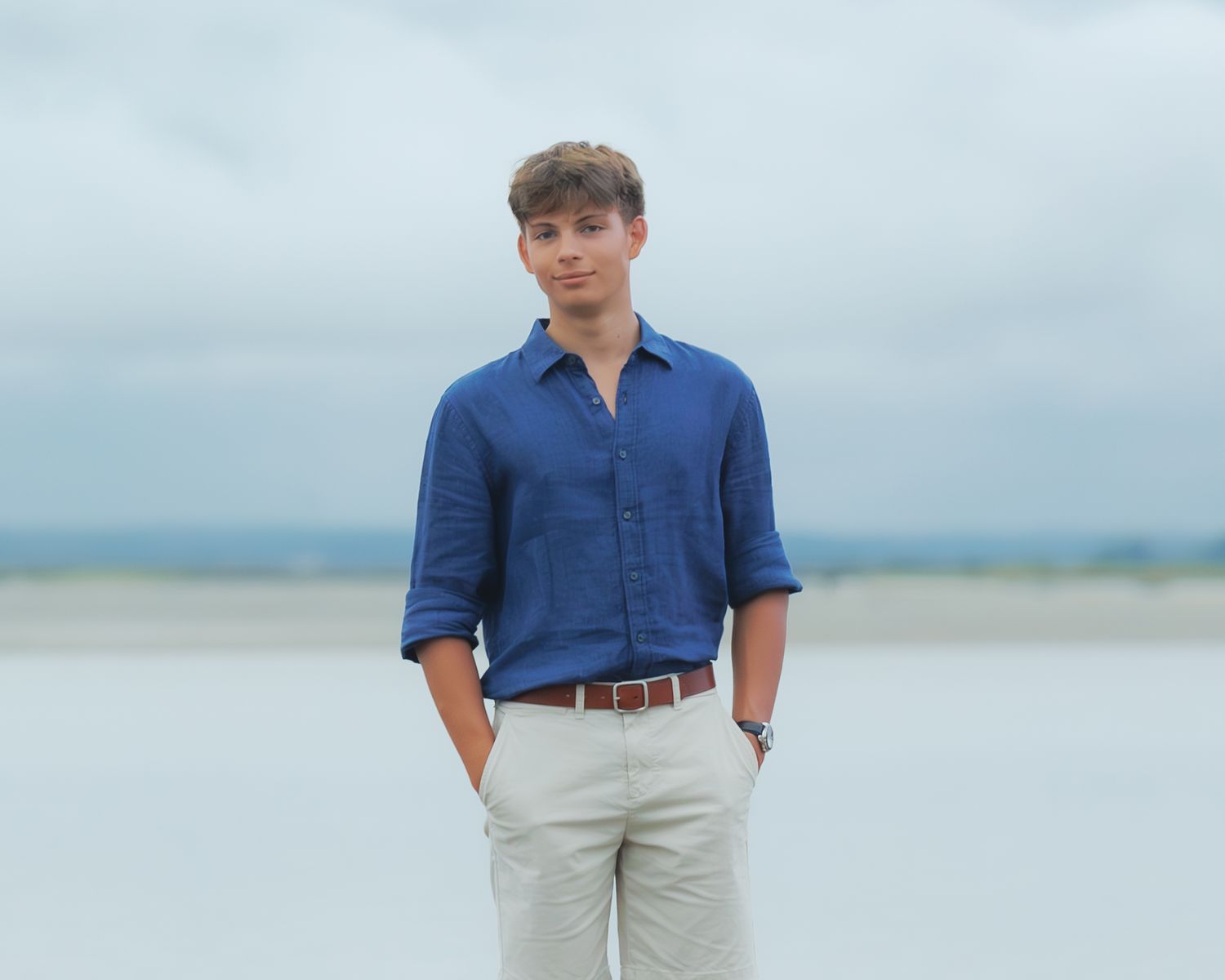 Young man in blue linen shirt and beige trousers standing on a white sand beach under cloudy skies.