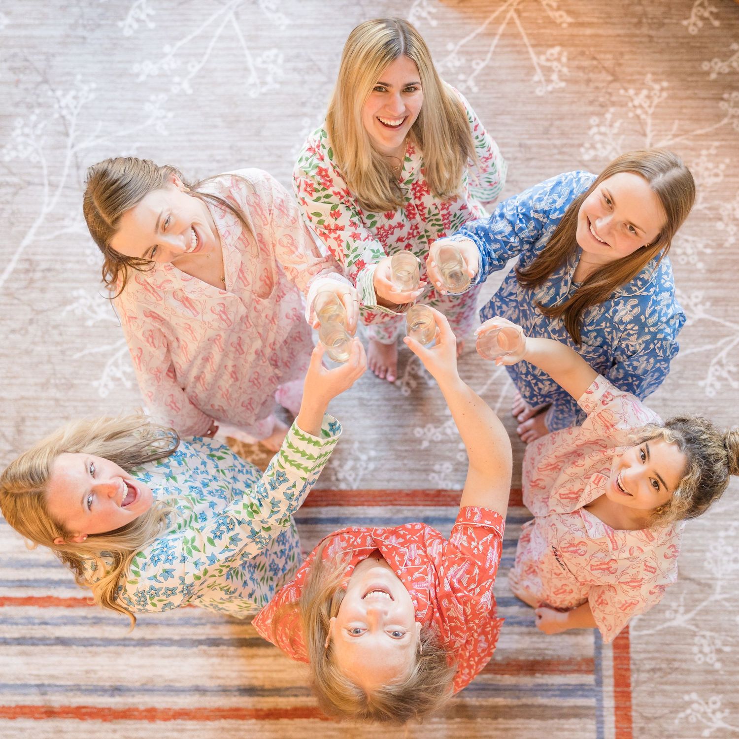 Six women in floral pajamas toasting drinks in a circle, viewed from above on a patterned rug.