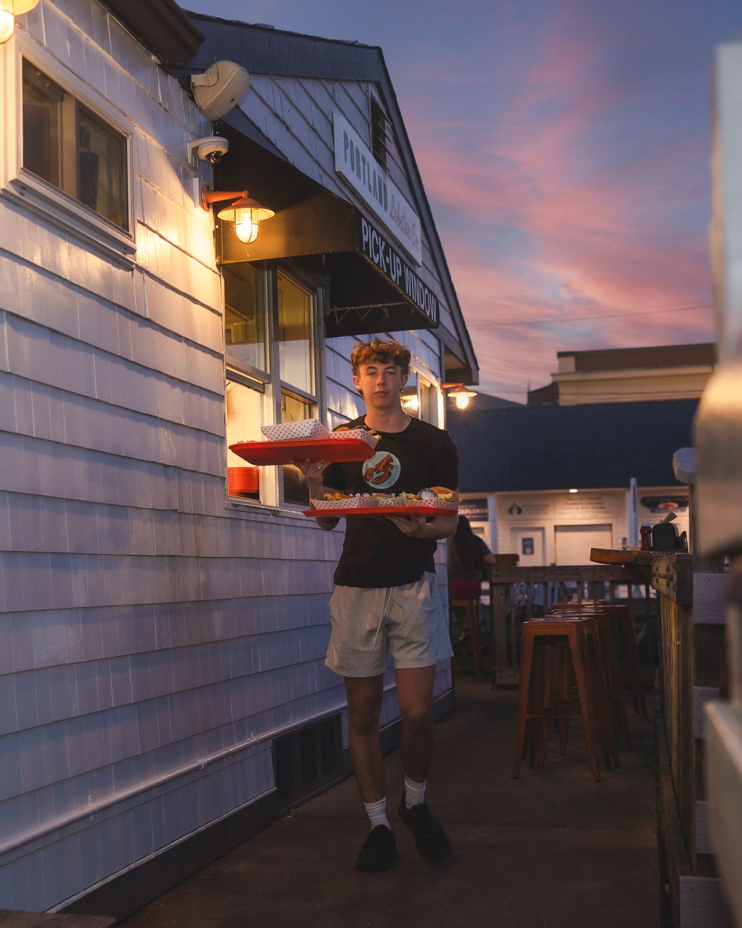 A young waiter carries a tray of food outside a coastal restaurant at sunset with a colorful pink sky backdrop.