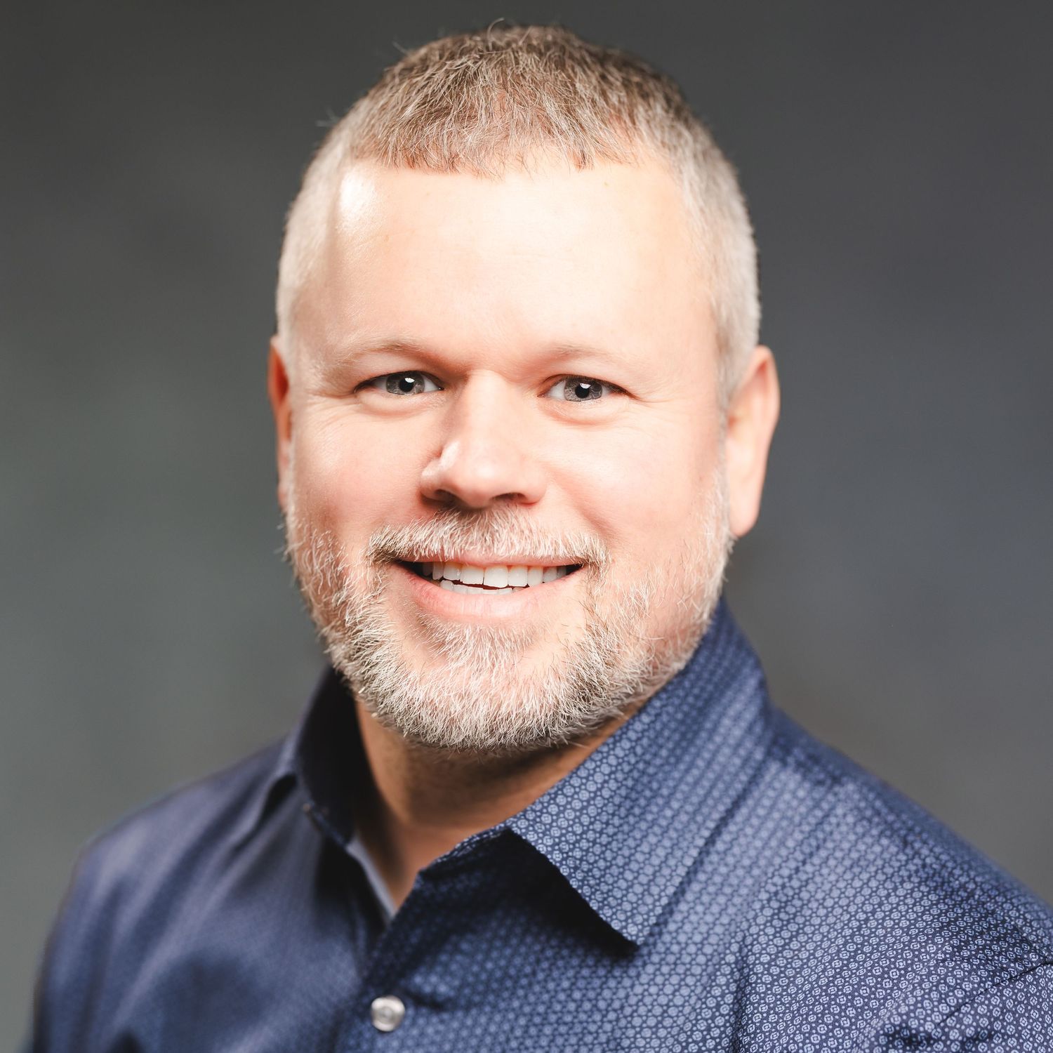 Southern Maine Headshot Photographer -Professional headshot of person with beard wearing blue collared shirt against dark background.