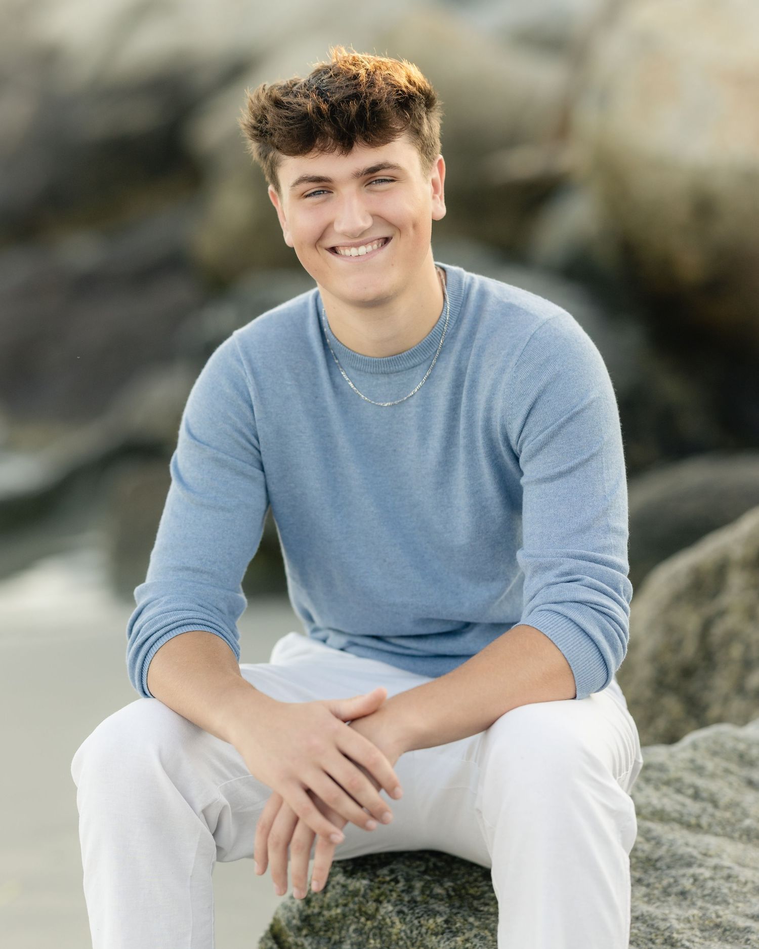 Smiling young man in blue sweater and white pants sitting on rocks at a beach for senior photos.