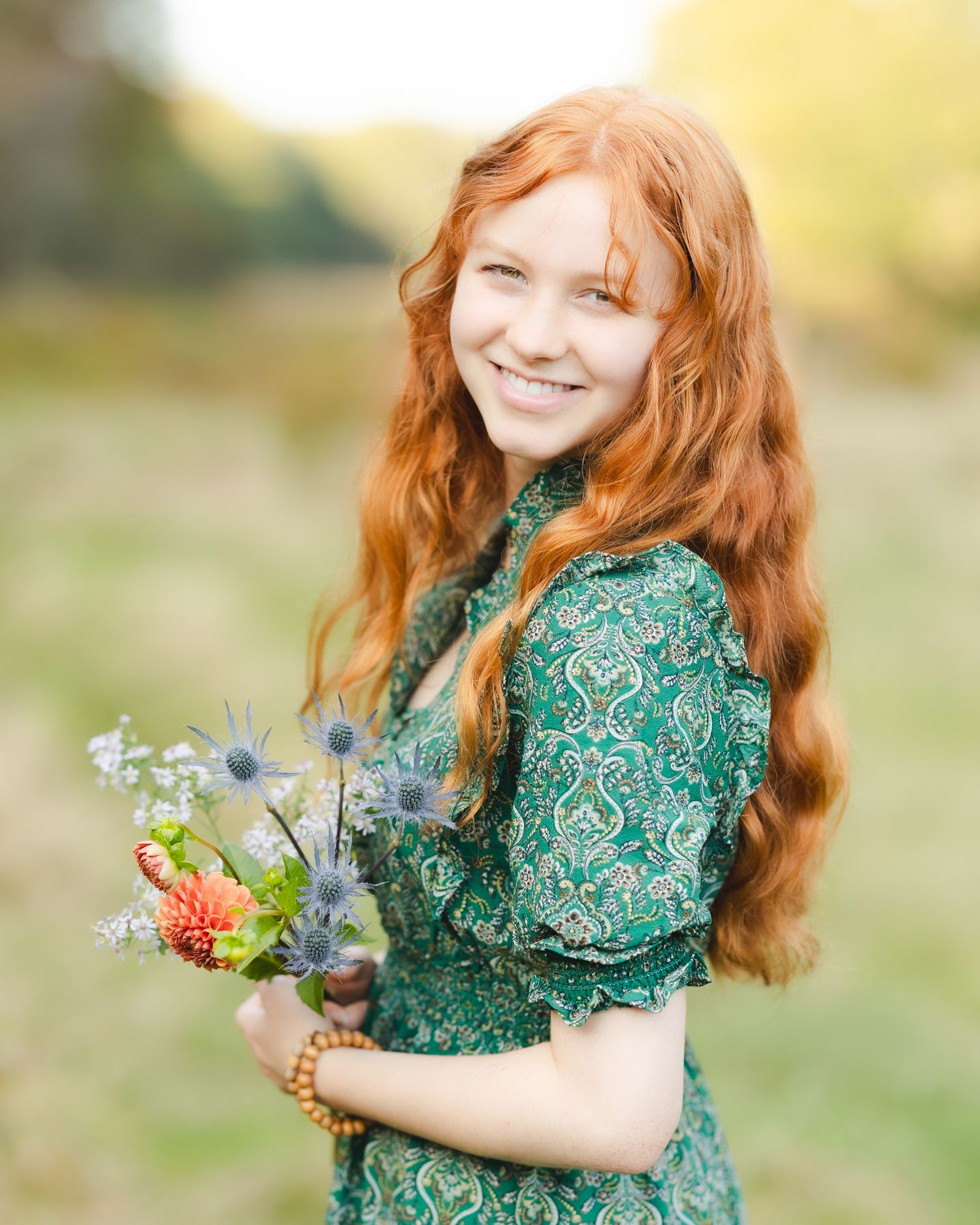 Smiling young woman with long red hair holding wildflowers, wearing a green floral dress outdoors.