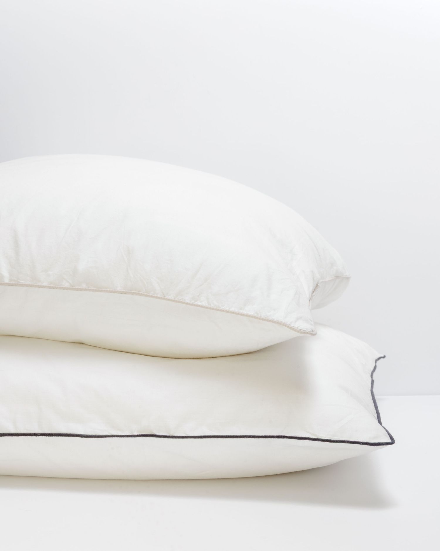 Stack of three white pillows with dark piping detail on a light surface, perfect for a luxurious bedding setup.