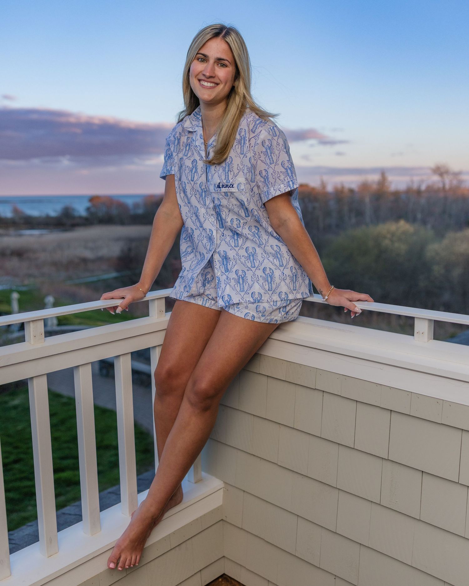 Woman in blue floral pajama set posing on a balcony railing at sunset with ocean views in background.