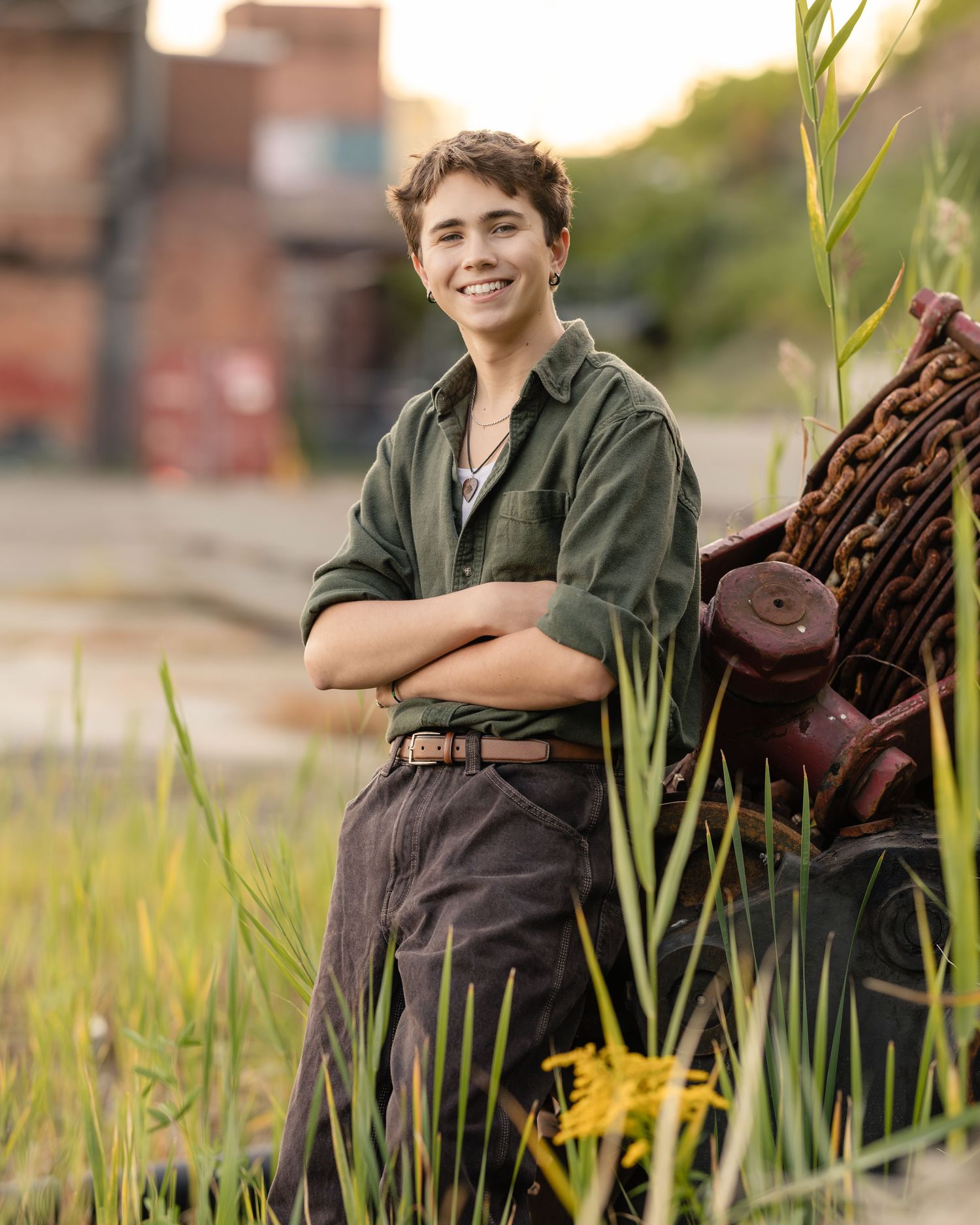 Smiling teenage boy in green shirt with crossed arms leaning against rustic machinery in a field at golden hour.