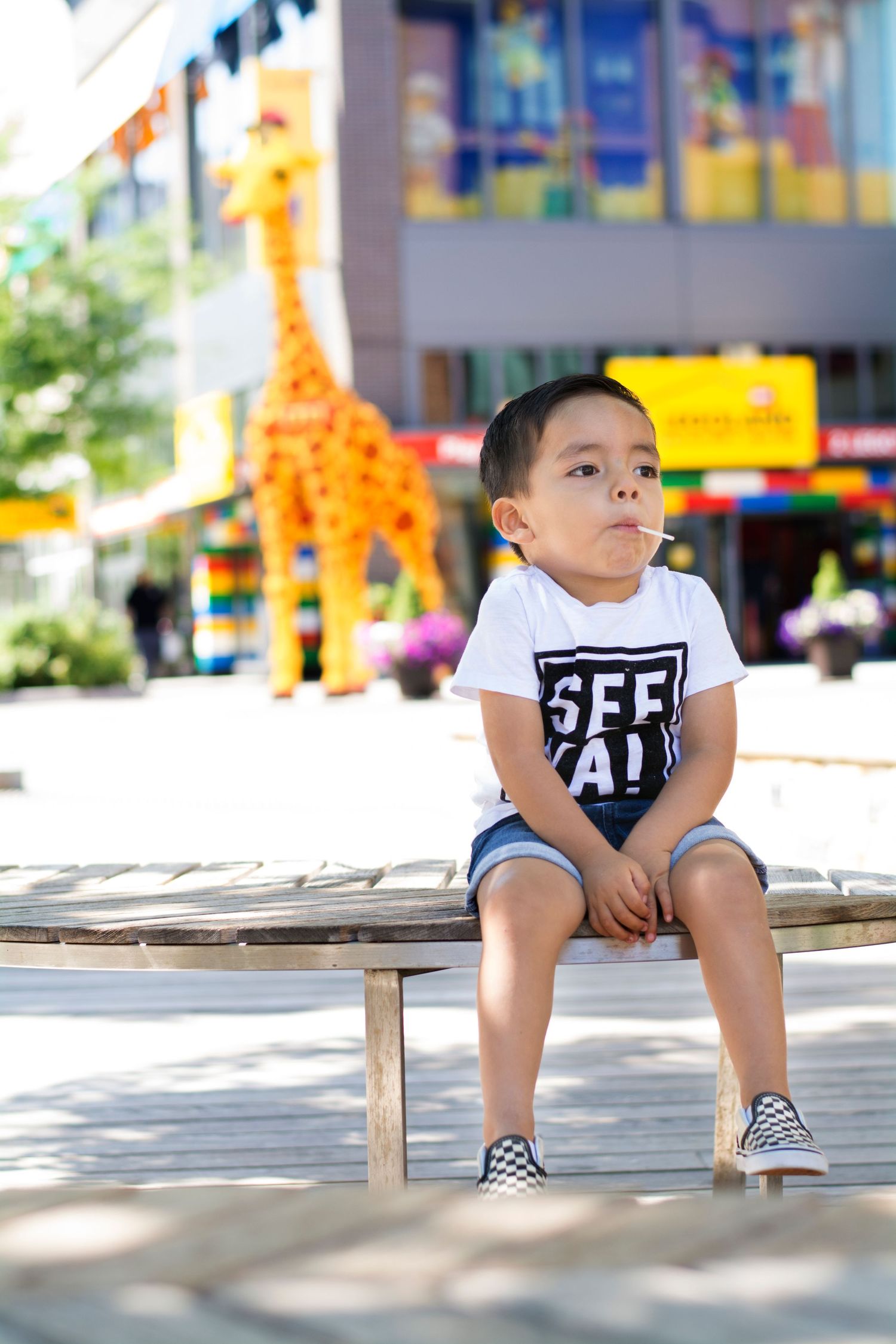 Child in BFF AT shirt sits on wooden bench with orange giraffe statue in background.