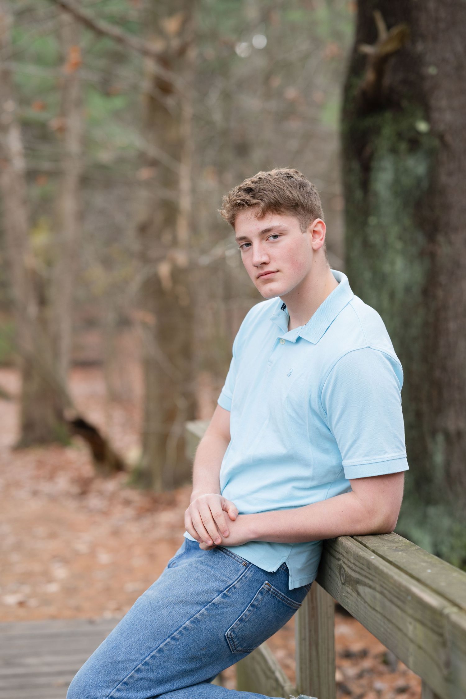 Young man in light blue polo shirt and jeans leaning on wooden railing in wooded outdoor setting.