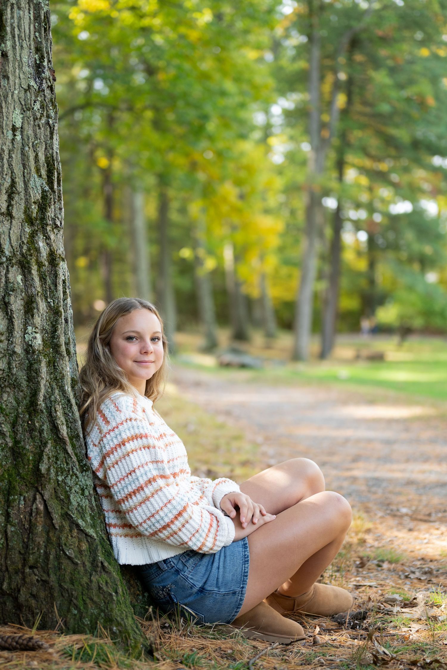 Young woman in striped sweater and denim shorts sits against a tree along a scenic autumn forest path.