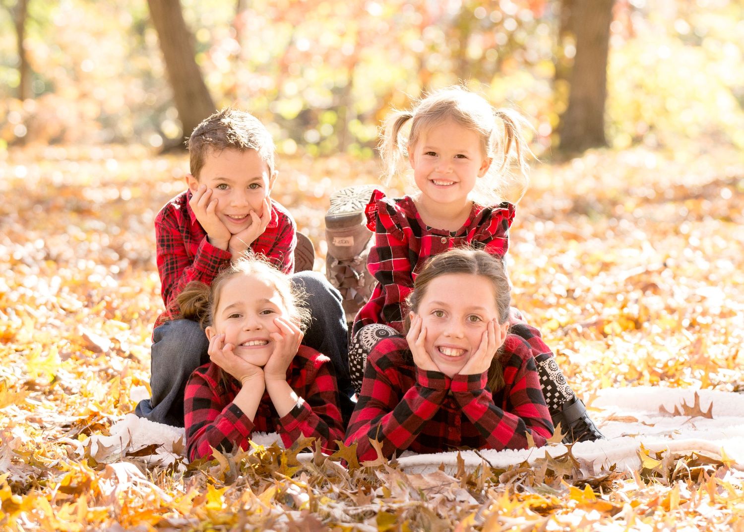 Four children in matching red plaid outfits lay together on autumn leaves during a fall photo session.