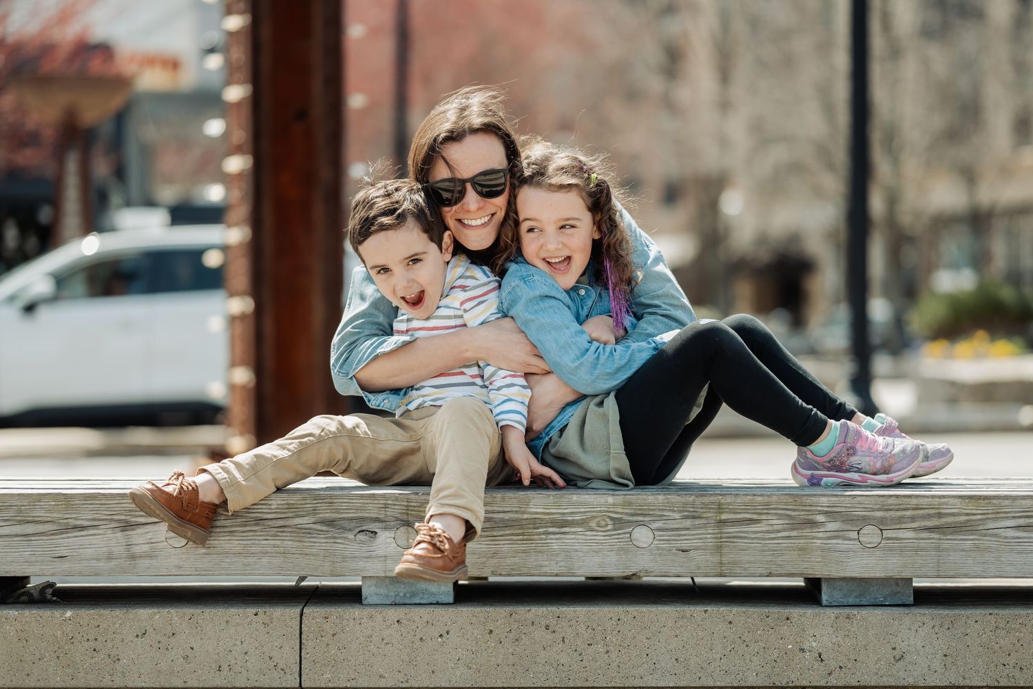 A smiling mother hugs her two young children while sitting outdoors on a sunny day.