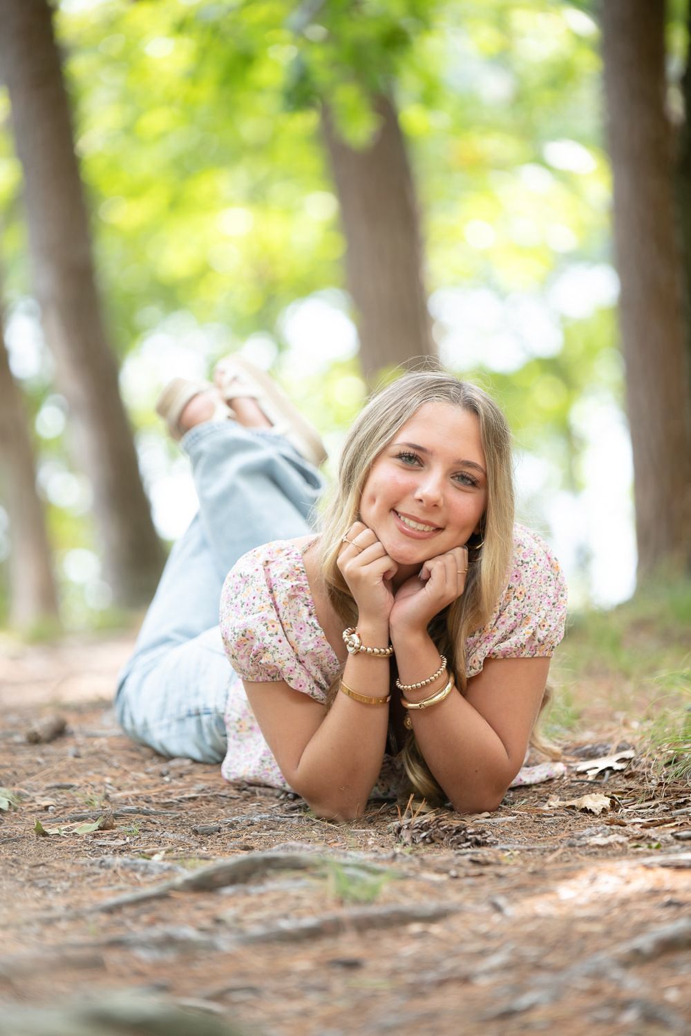 Young blonde woman lying on forest path, smiling with hands under chin, wearing floral top and jeans.