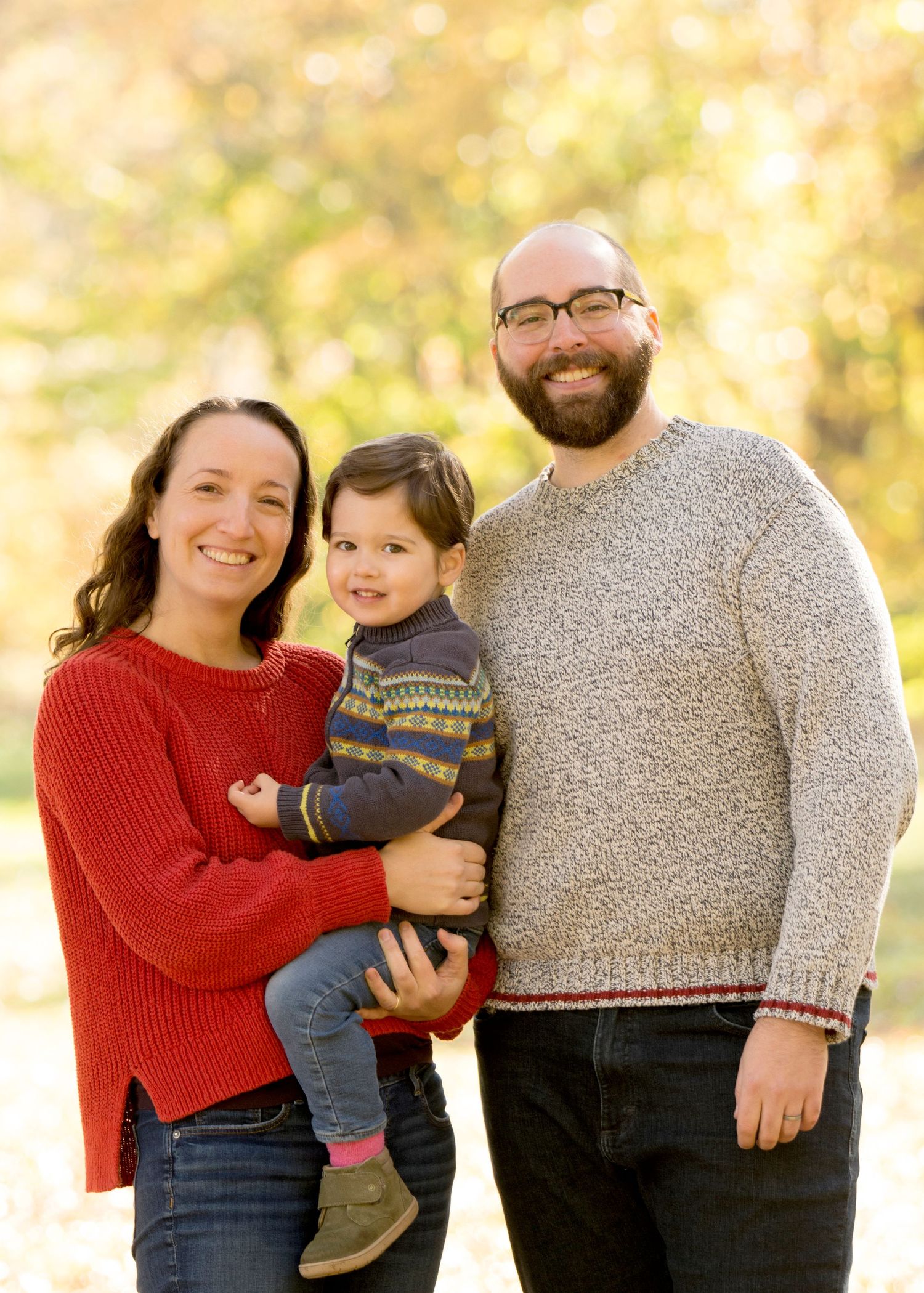 Family of three posing together in cozy sweaters against golden fall backdrop.