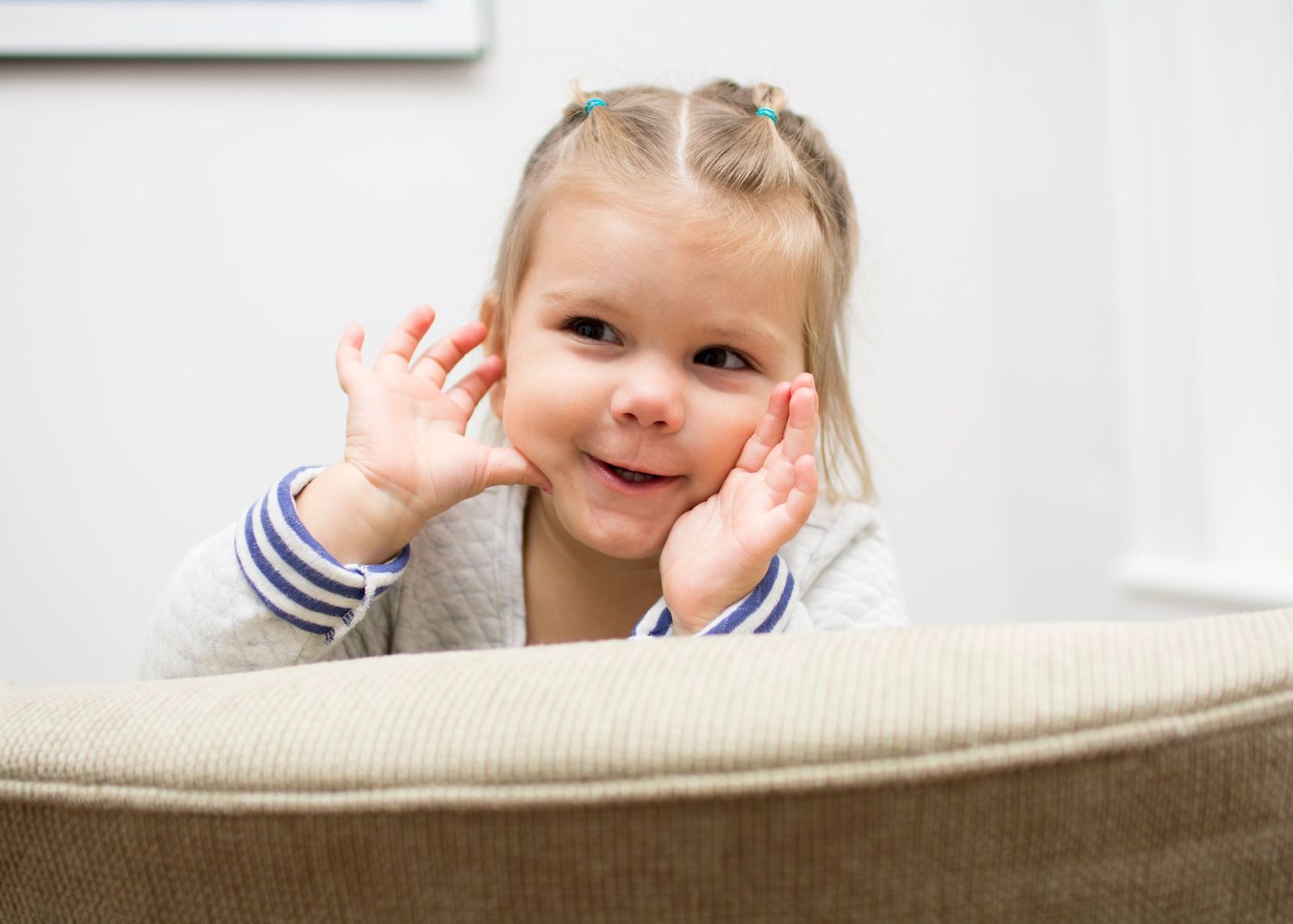 A child leans over a couch cushion with hands near their face in a playful pose.