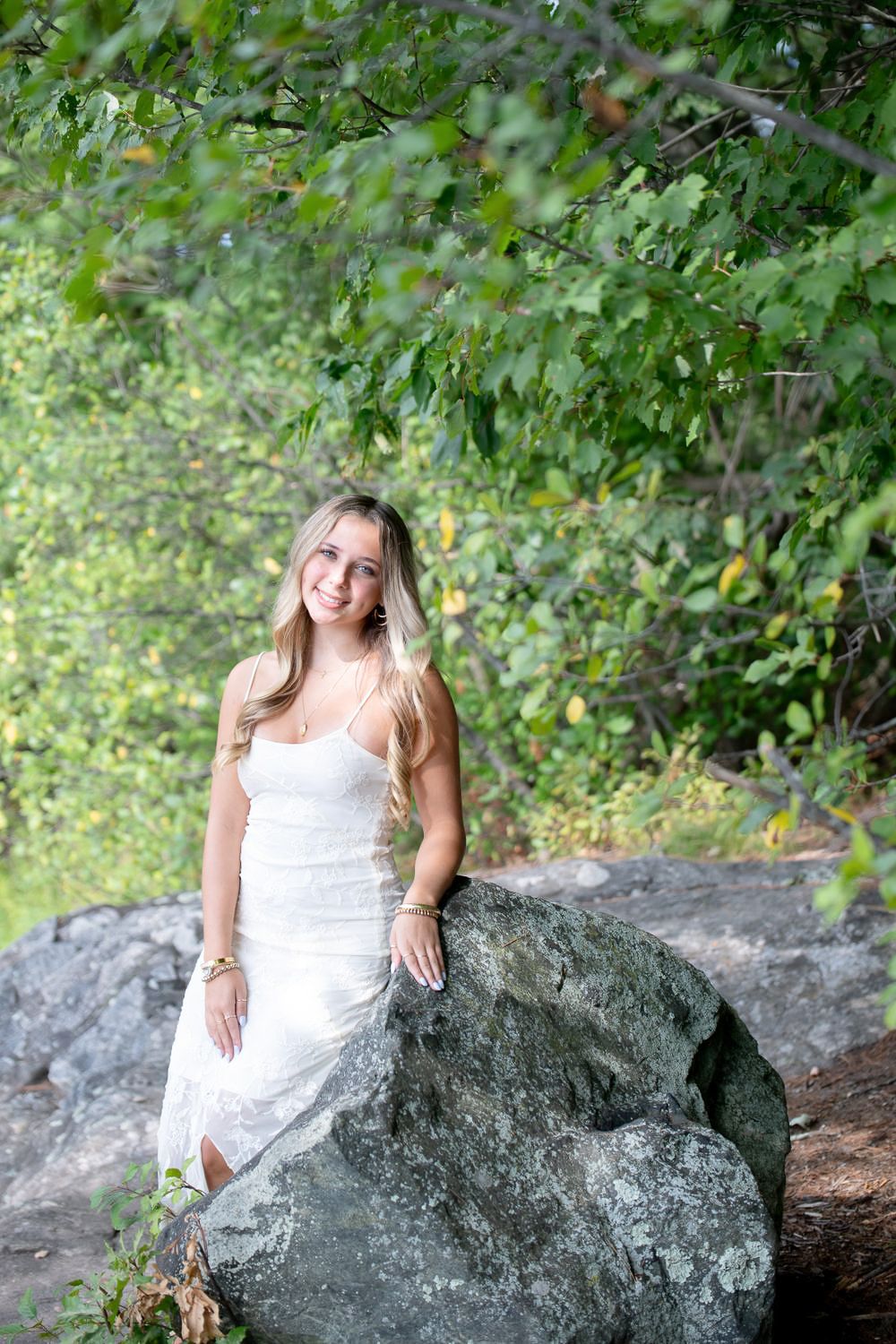 Young blonde woman in white dress sitting on a large rock surrounded by lush green trees outdoors.