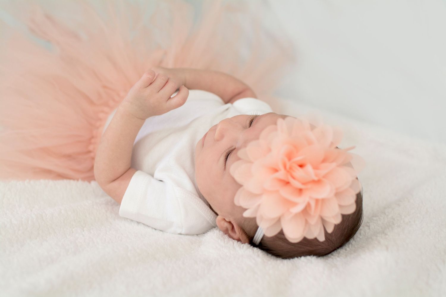 Baby in white outfit with peach flower headband sleeping on light backdrop.