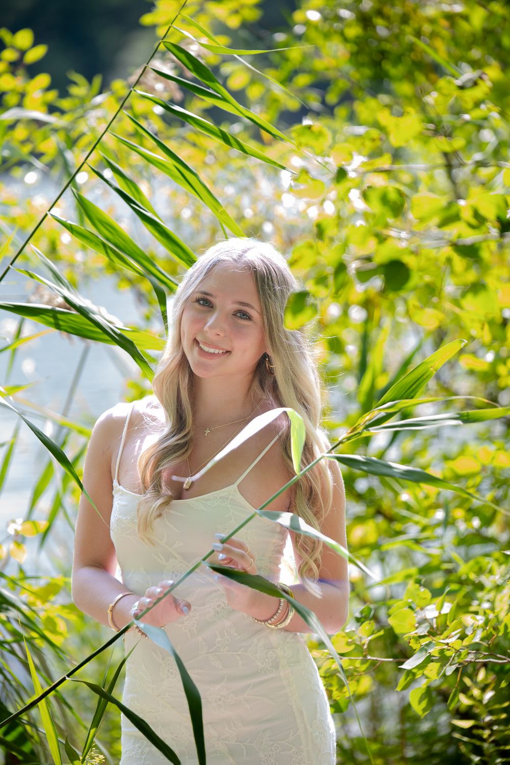 Smiling blonde woman in white dress surrounded by lush green foliage and tall grass in sunny outdoor setting.