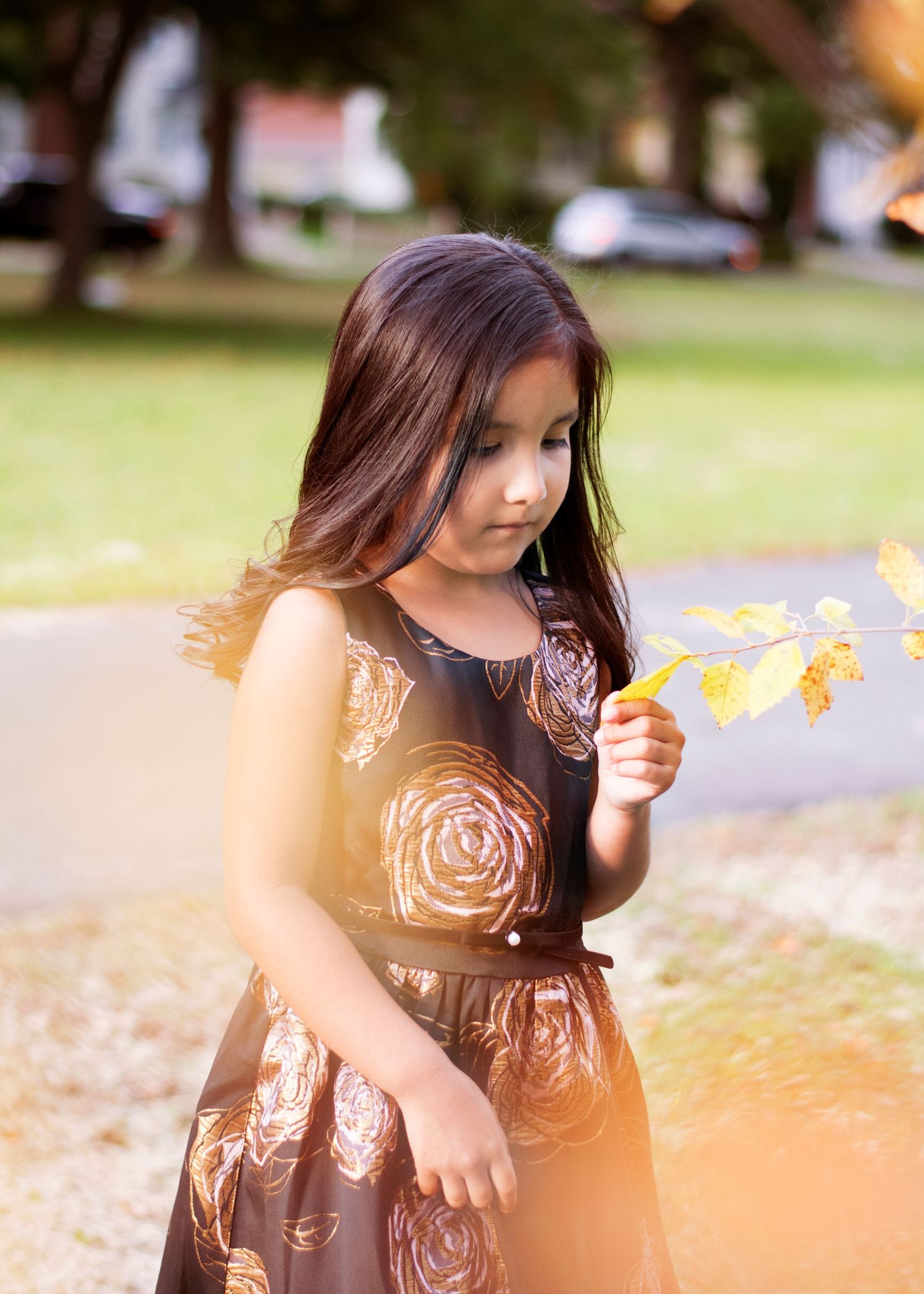 Child in brown floral dress holds autumn leaf while standing outdoors.