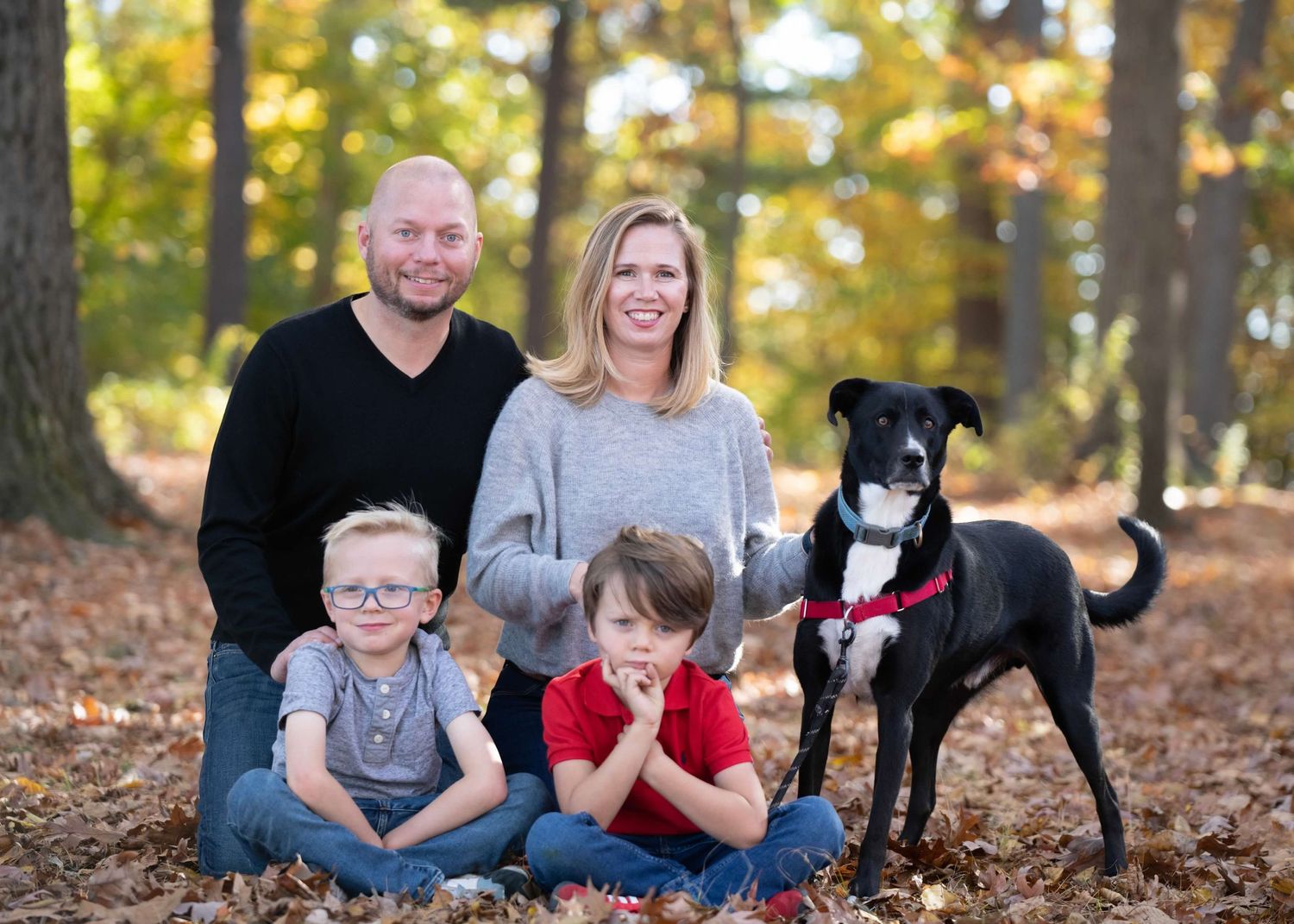 A family with their black and white dog poses together in a wooded autumn setting.