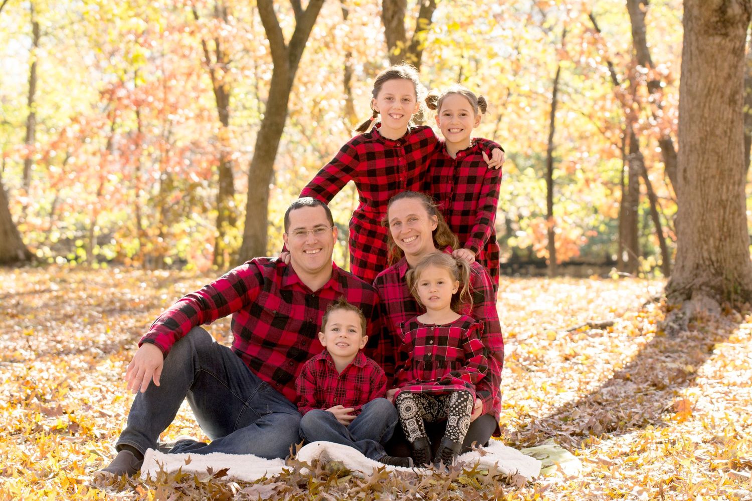 Family in matching buffalo plaid shirts posing together on ground in fall forest setting.