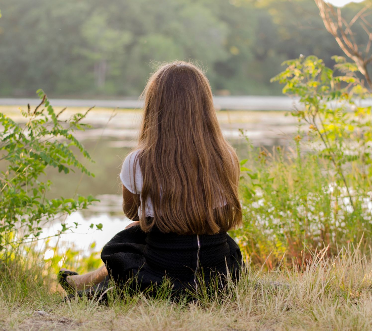 Person with long brown hair sits peacefully by a tranquil lake at sunset.