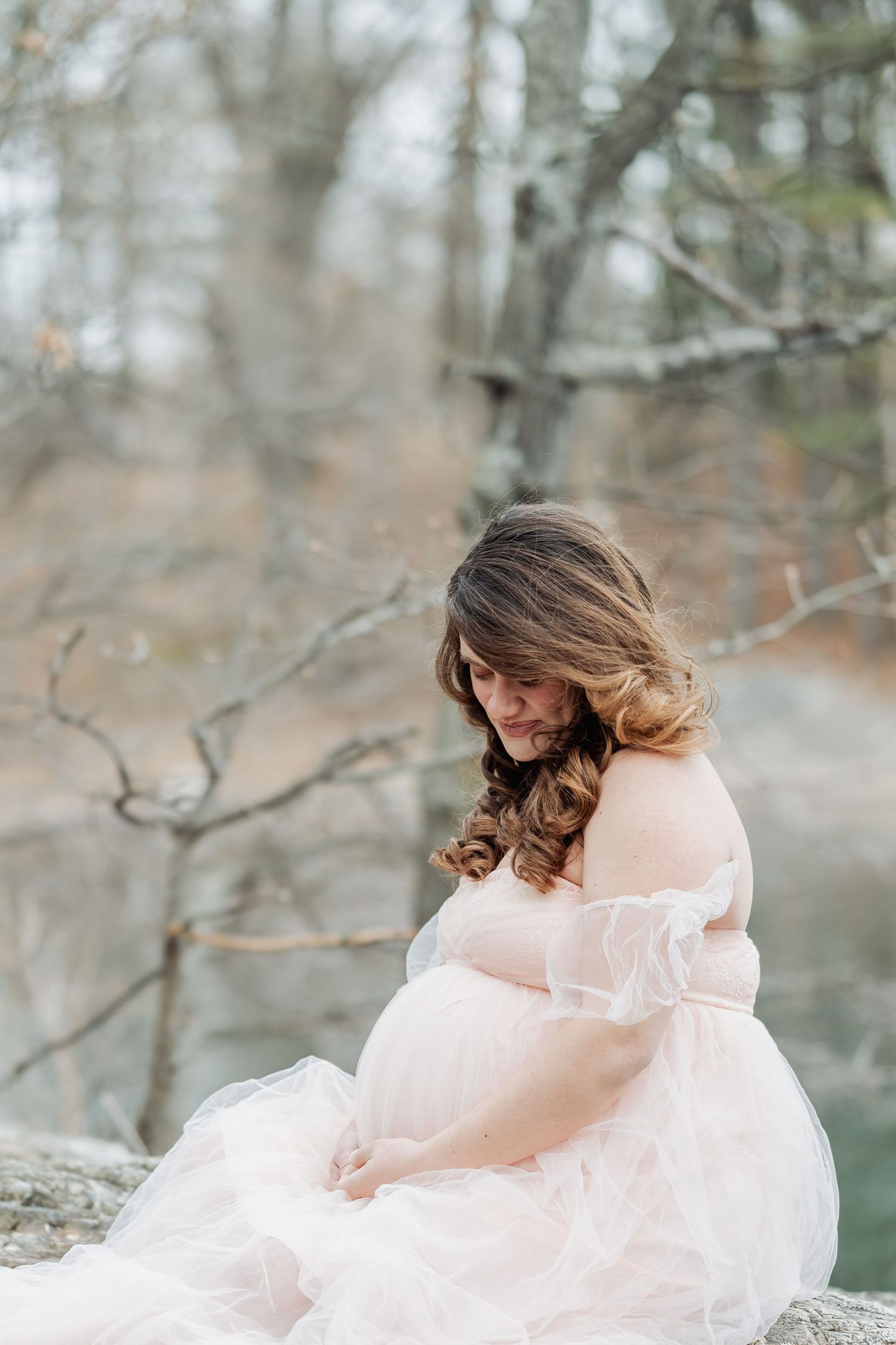 Pregnant woman in sheer blush gown sitting outdoors among bare trees, cradling her baby bump in a serene maternity portrait.