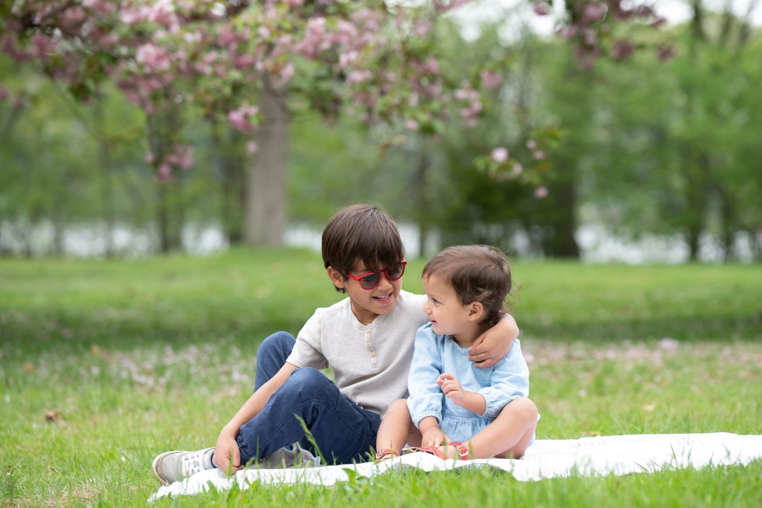 Two children sit together on a blanket in a spring park with blooming cherry trees in the background.