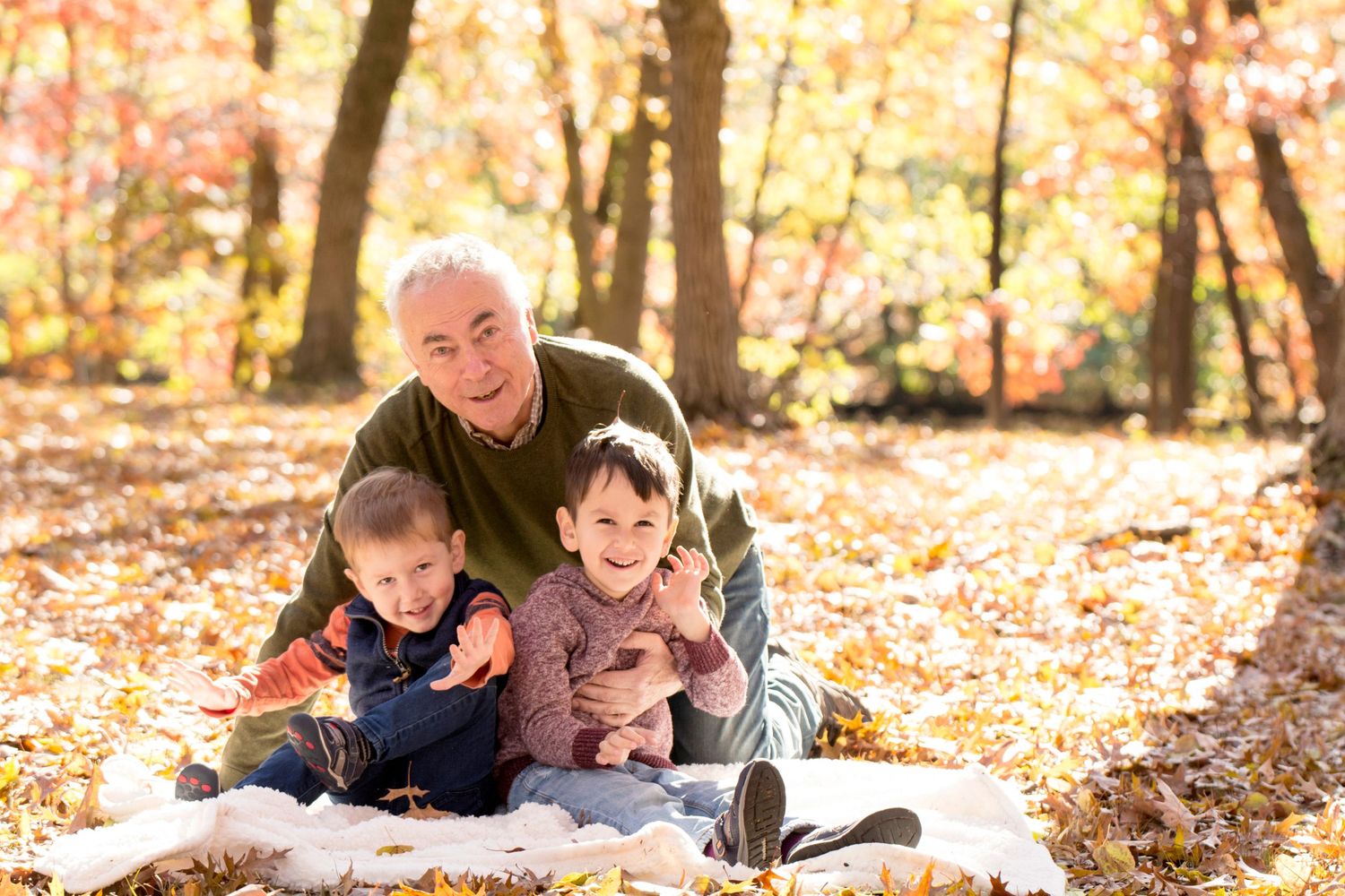 Three children sitting and smiling on white blanket in autumn forest setting.