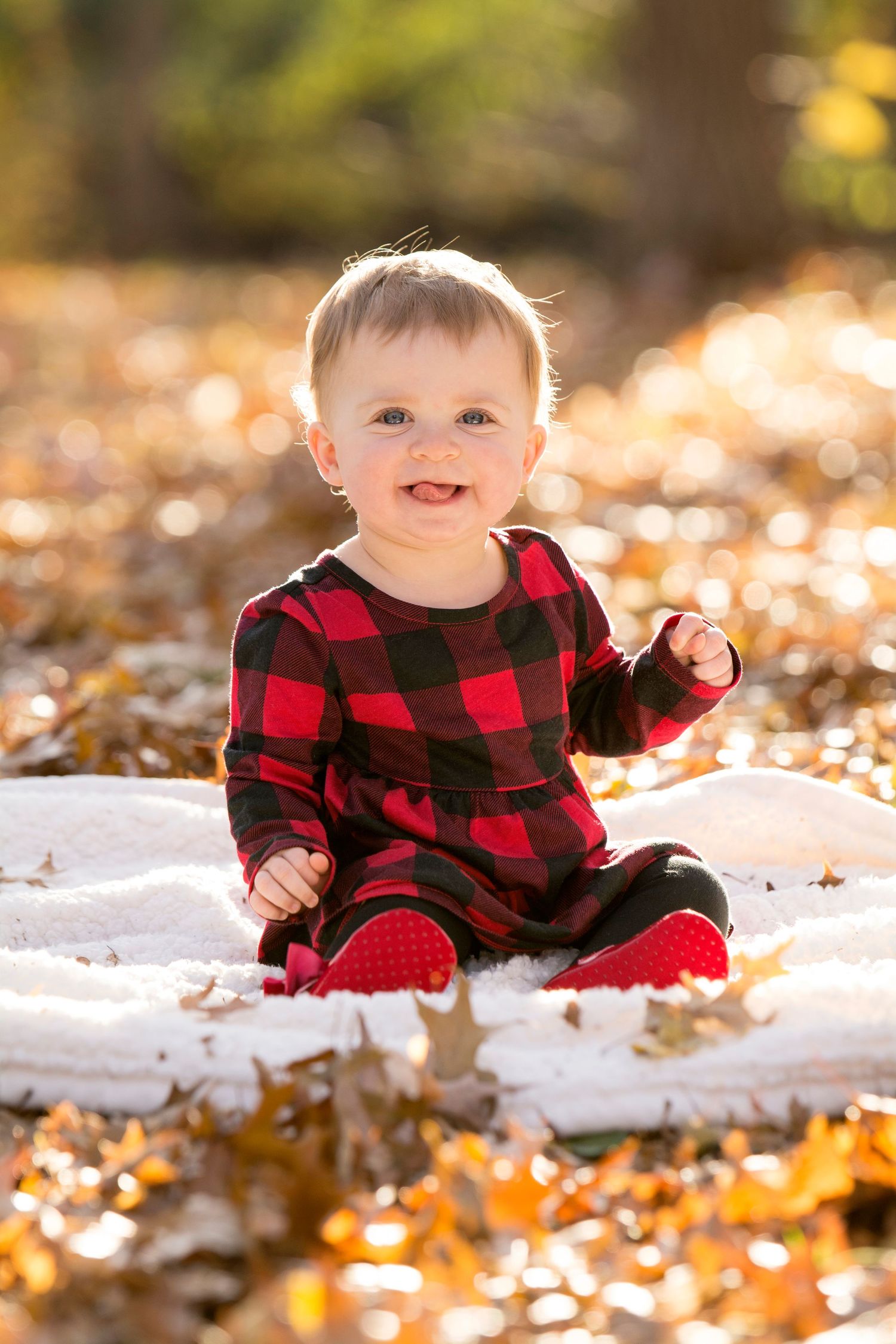 Baby wearing red buffalo plaid outfit sitting on white blanket among autumn leaves.