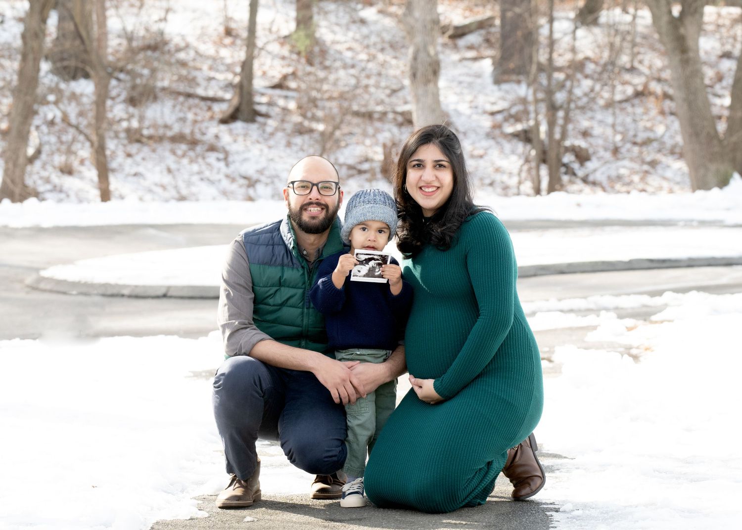Family of three poses together in winter scene with snow covered trees.