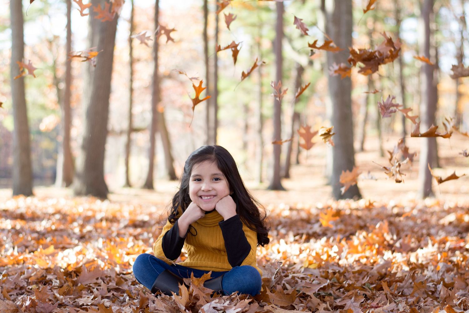 Child sitting cross-legged among fallen autumn leaves in wooded setting.