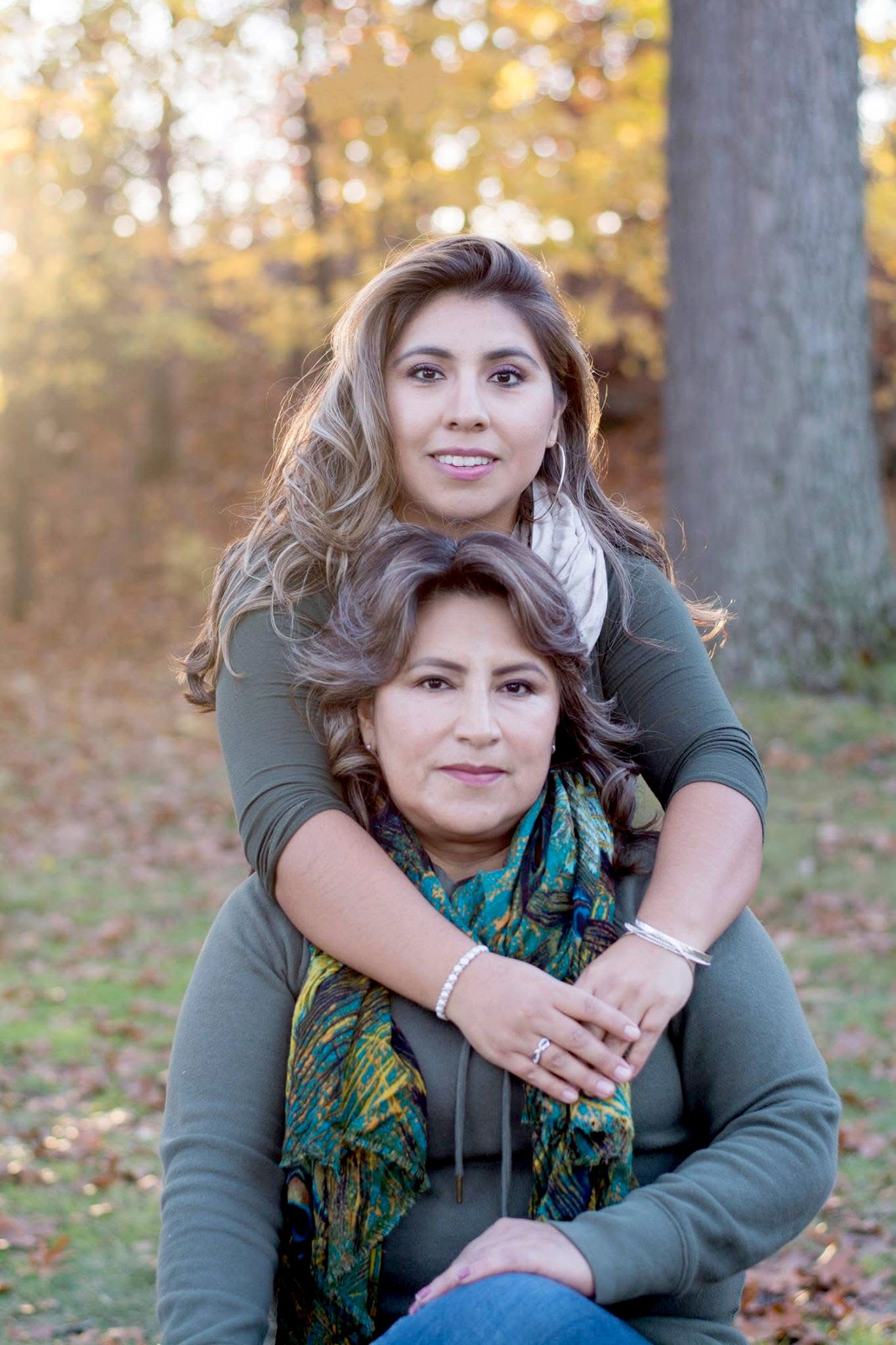 Two people embracing outdoors against autumn foliage wearing green sweaters and patterned scarf.