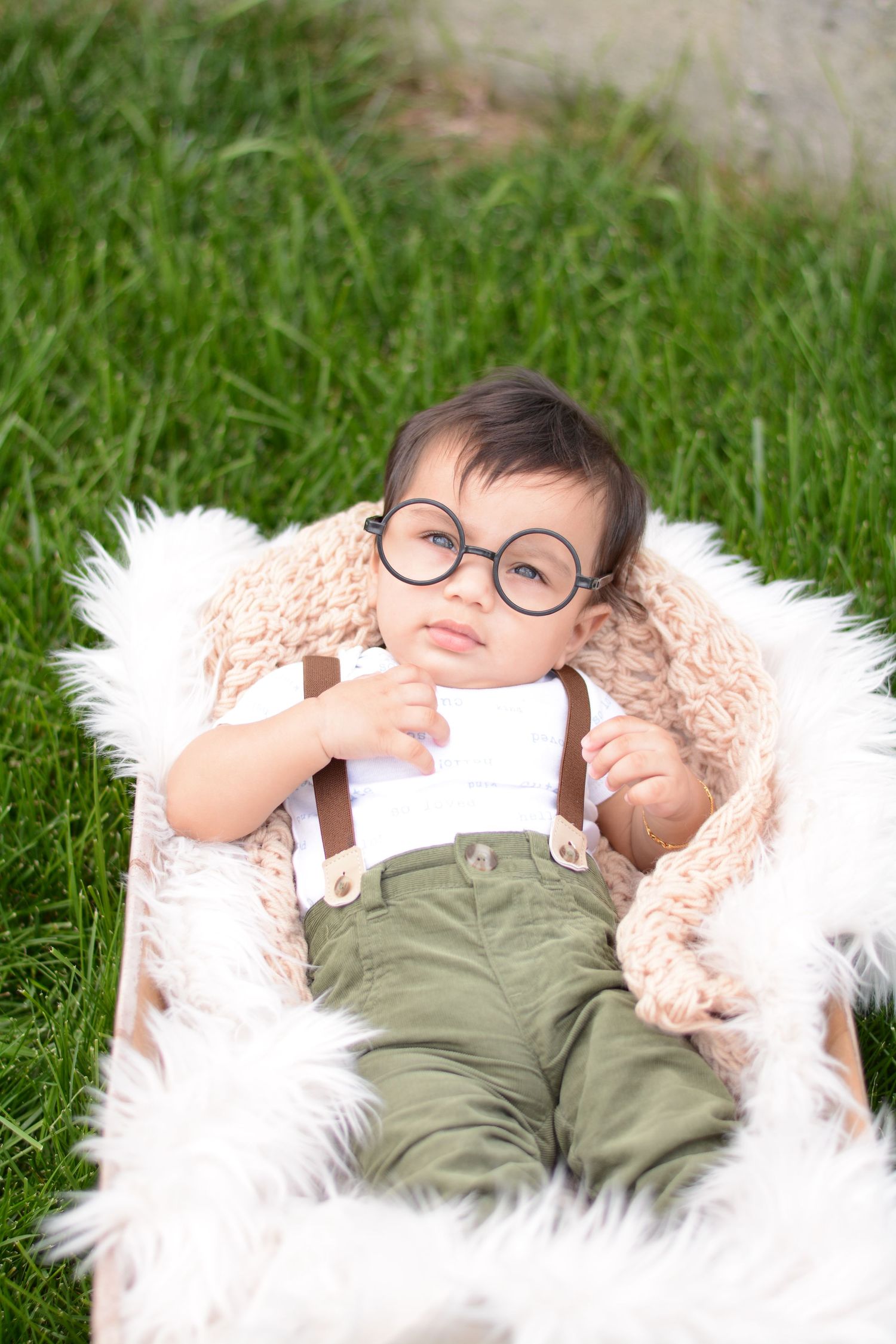 Baby wearing glasses and overalls reclines on fluffy white blanket in grass.