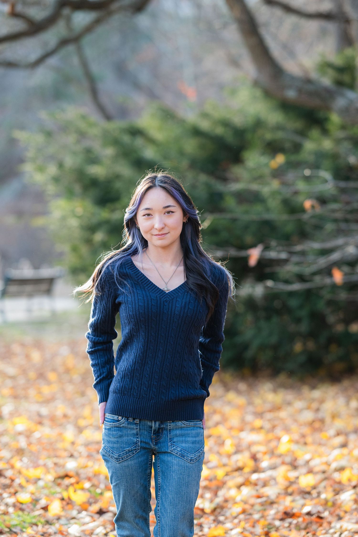 Young Asian woman in navy sweater and jeans standing among autumn leaves in a park.