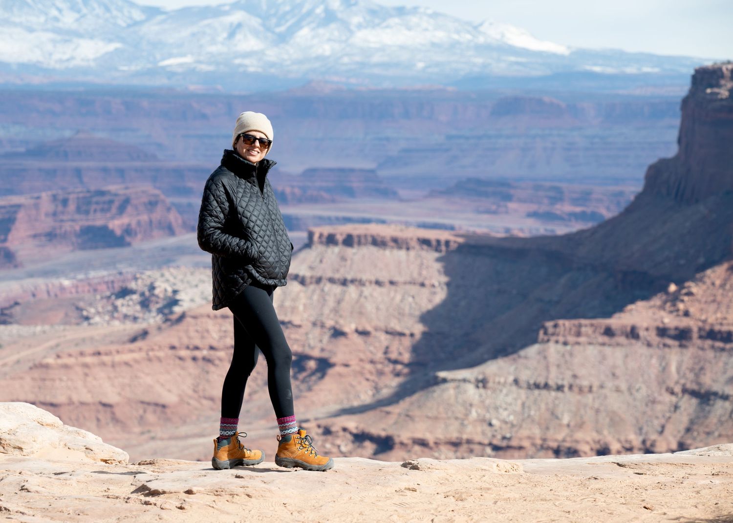 Person in winter jacket stands on cliff edge overlooking vast canyon landscapes.