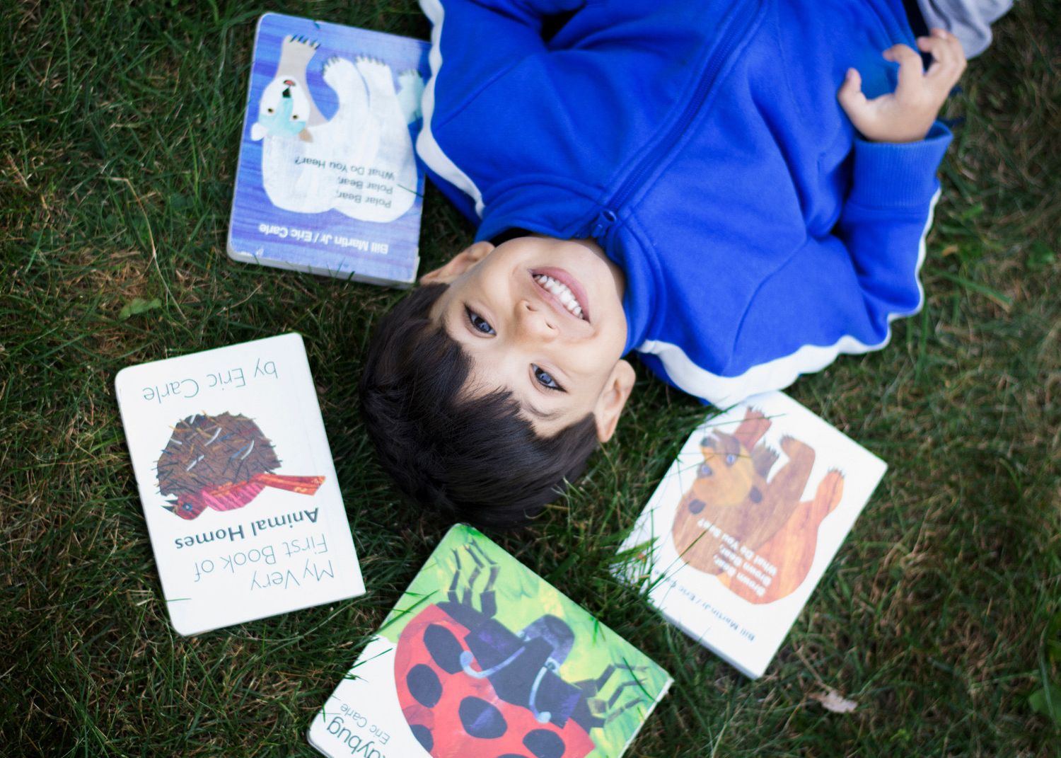 A child lying on grass surrounded by children's picture books spread out around them.