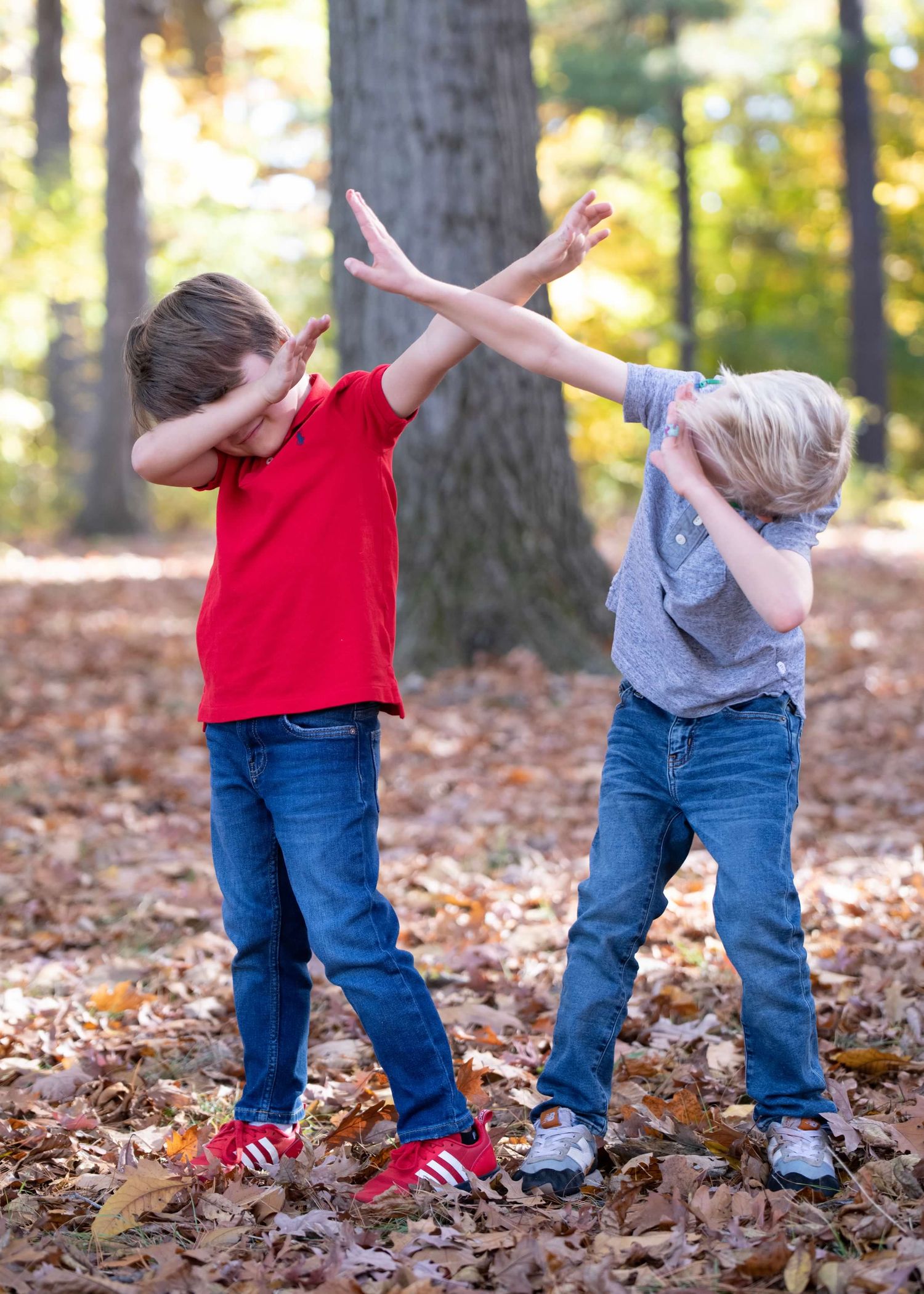 Two young boys in casual wear doing dab dance moves among fallen leaves in forest.