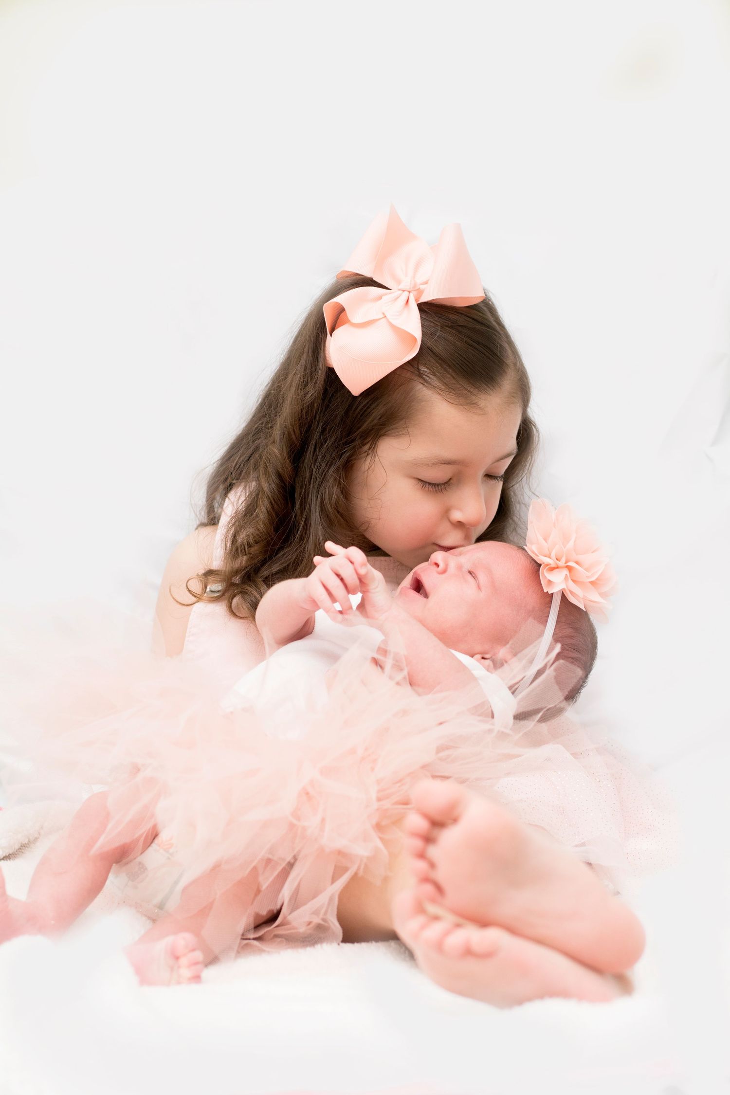 Two siblings in soft pink tulle outfits share a tender moment on white backdrop.