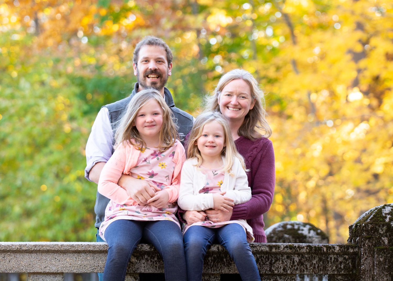 Family of four sitting together outdoors with autumn foliage in background.