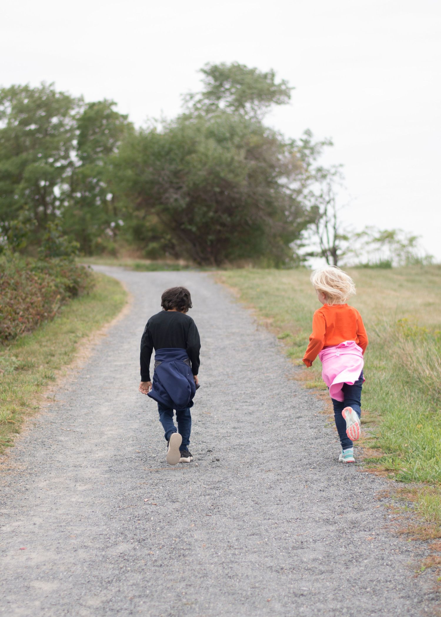Two children running together on a gravel path surrounded by green trees and grass on a sunny day.