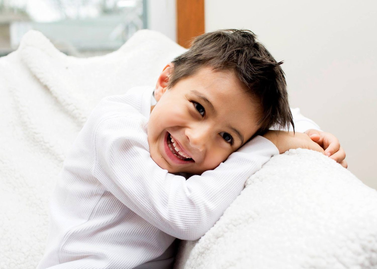 A young child in white lounges comfortably on white bedding with a bright smile.