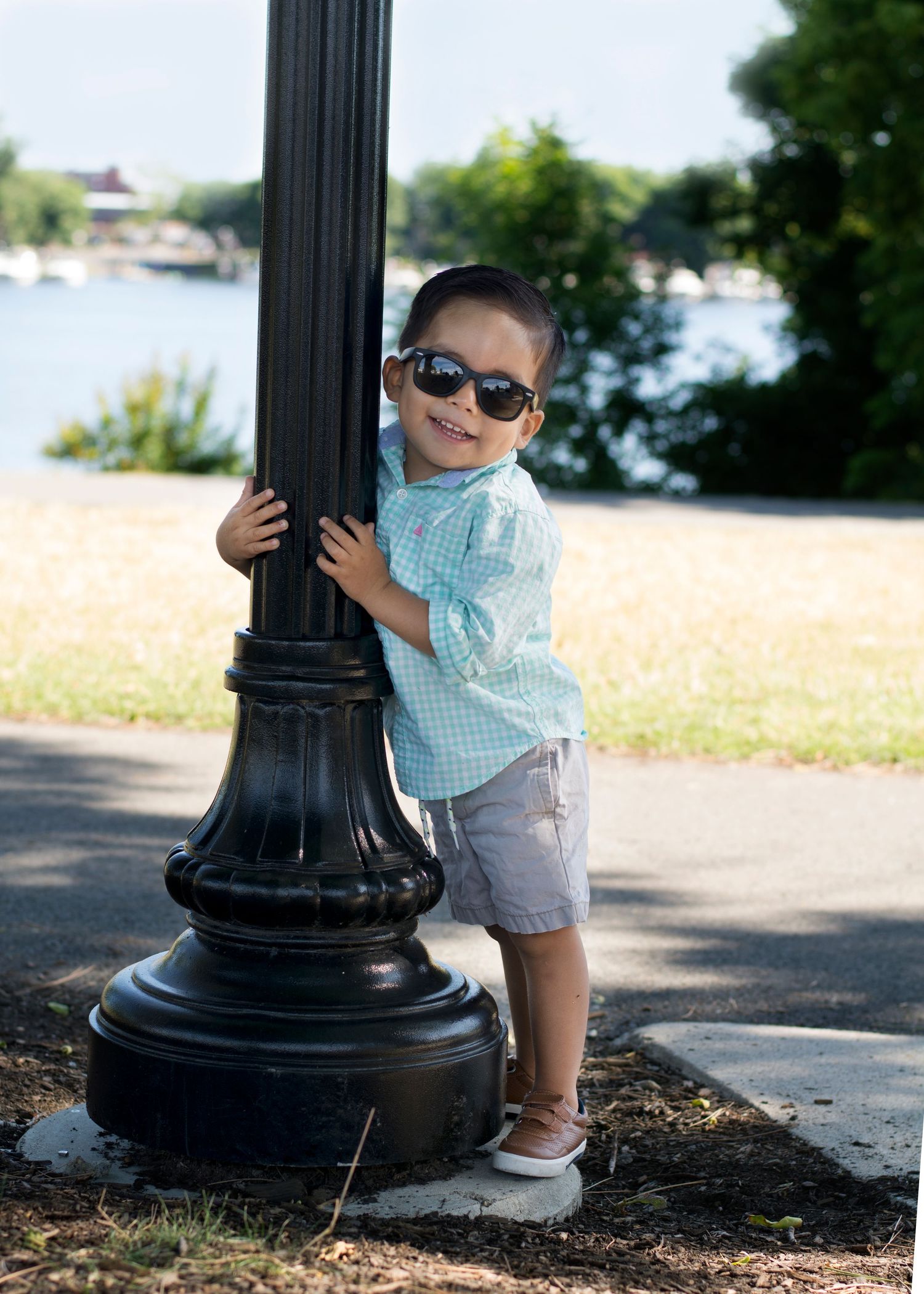 Small child in checkered shirt and sunglasses poses next to black lamp post near waterfront.